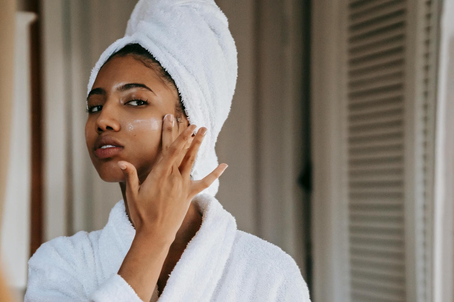 Young ethnic female in robe with terry towel on head applying moisturizing cream on face while looking in mirror in house - best natural moisturizer