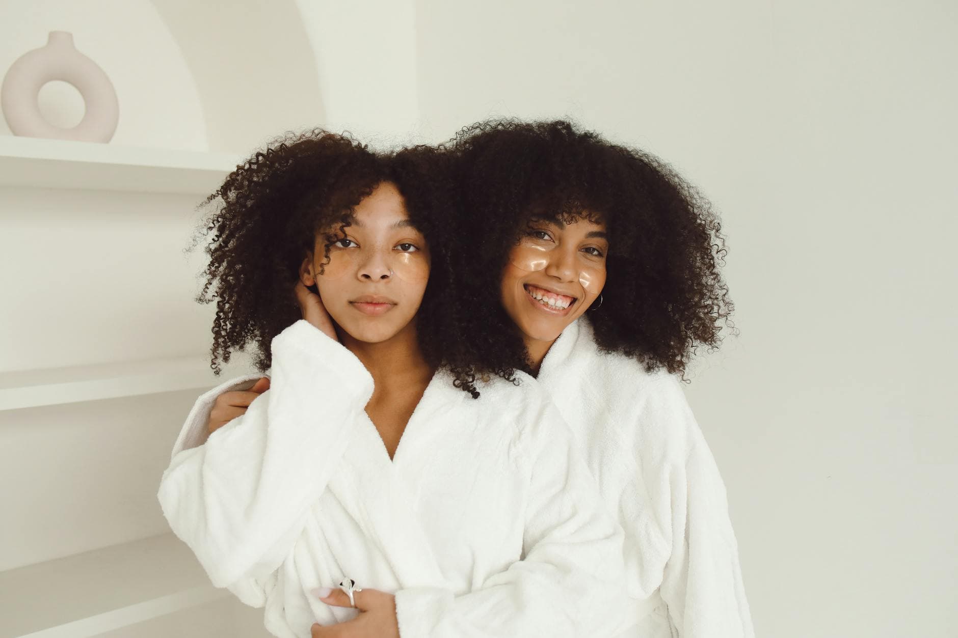 Two women enjoying a skincare routine with facial patches and bathrobes, embracing each other joyfully indoors. - best natural skin care