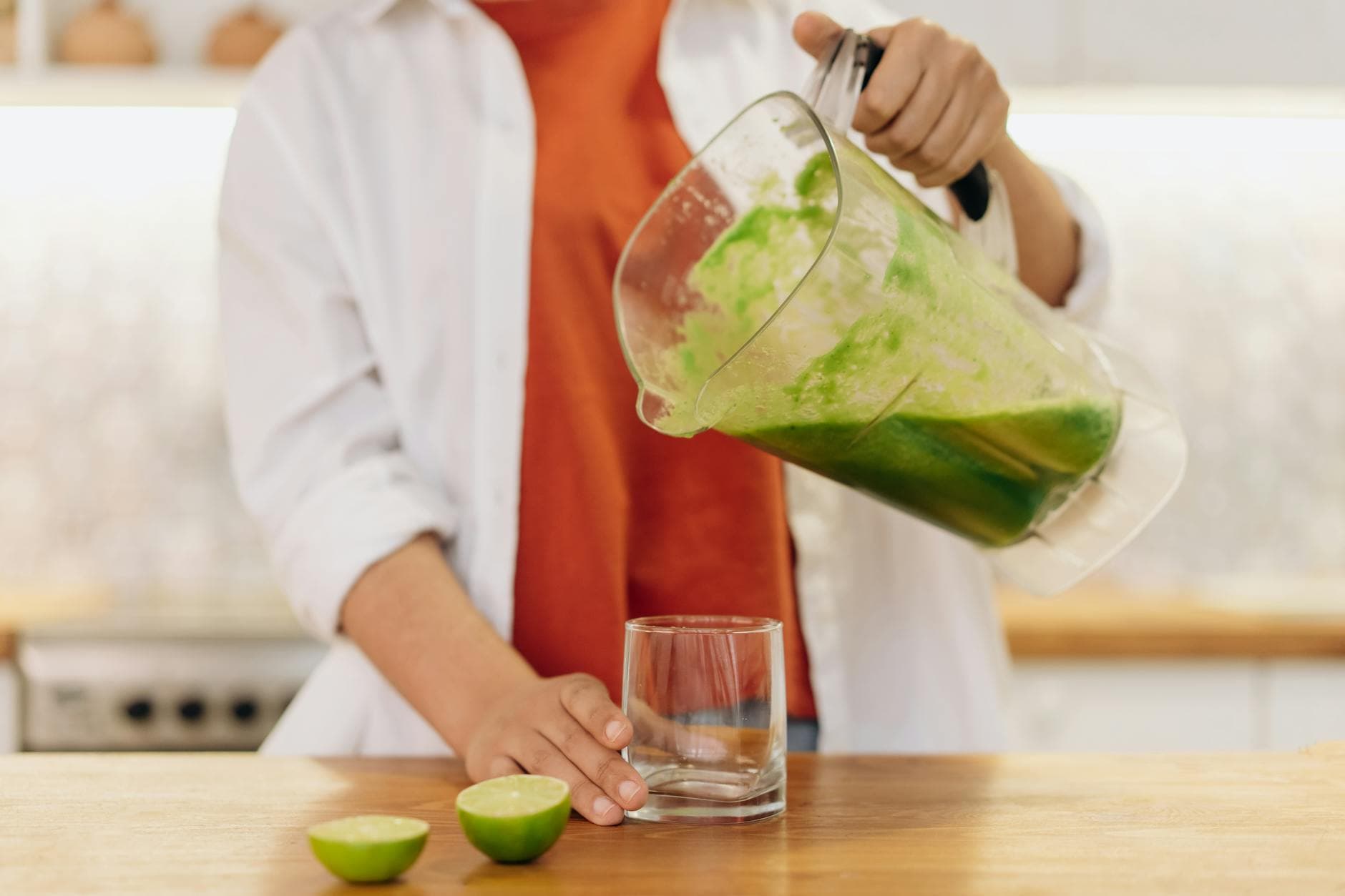 Close-up of a person pouring a vibrant green smoothie into a glass with limes on the wooden table. - body detox cleanse