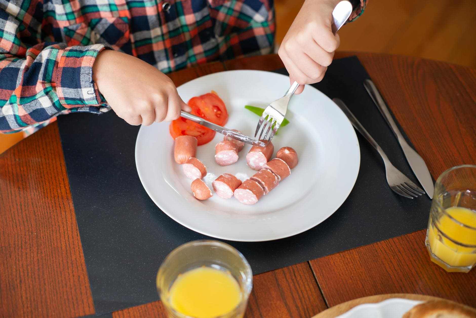 A child slices sausages at a breakfast table with juice, showing a cozy morning meal scene. - healthy morning habits