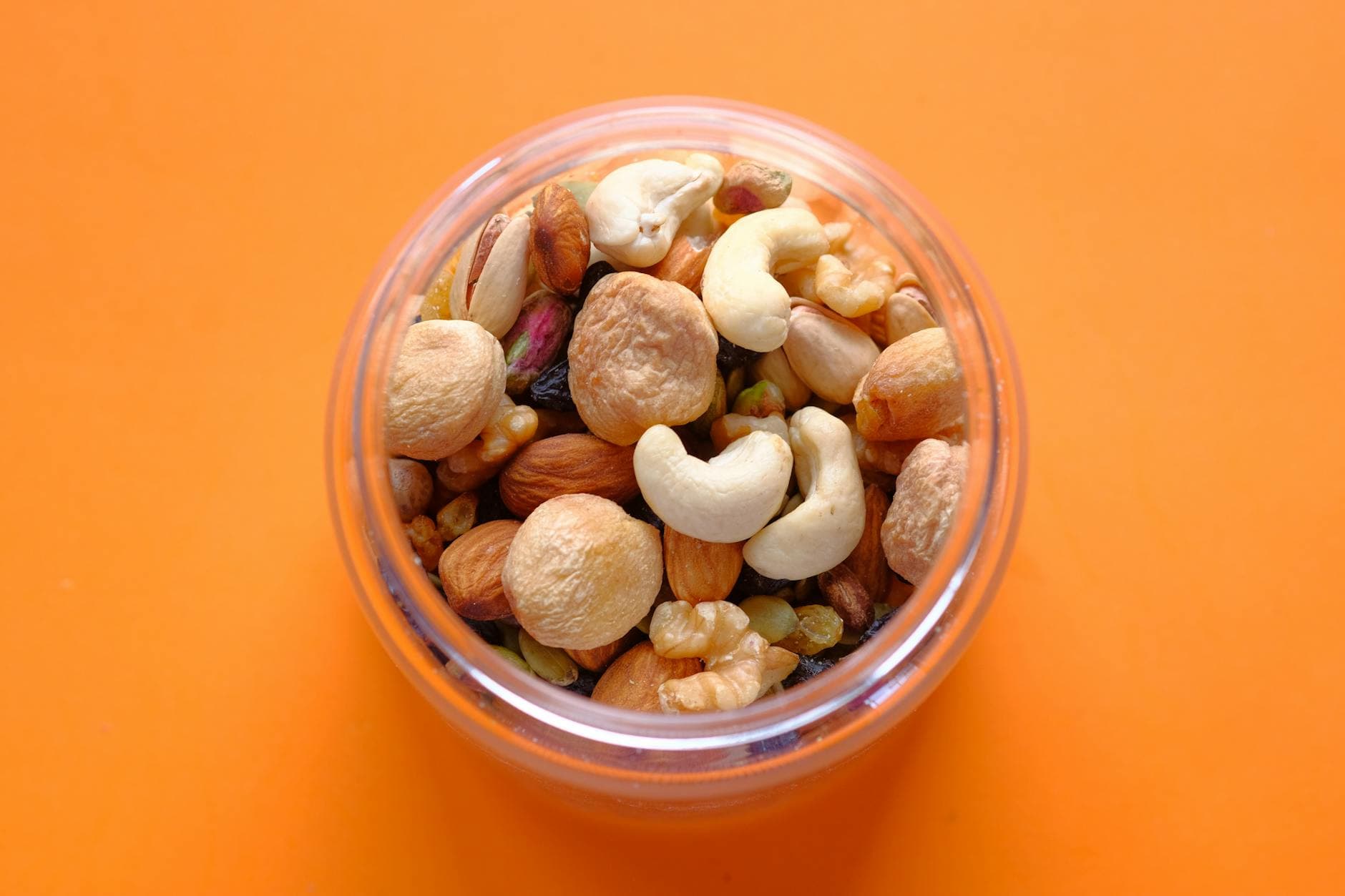 Close-up of a mix of nuts in a clear container against an orange background. - healthy office snacks