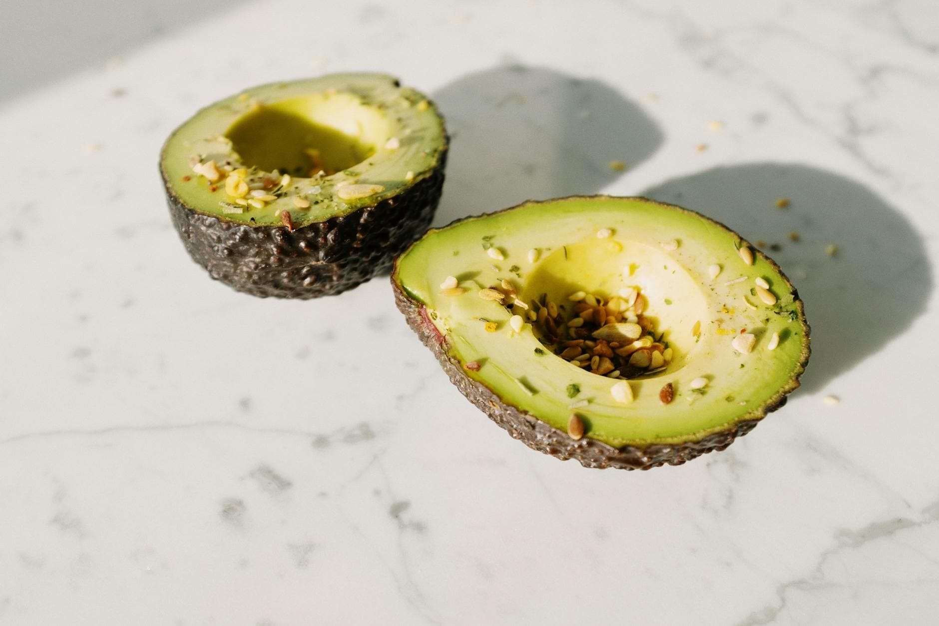 From above of green halved avocado without kernel sprinkled with various seeds placed on white marble desk served for healthy vegan breakfast preparation - healthy spring snacks