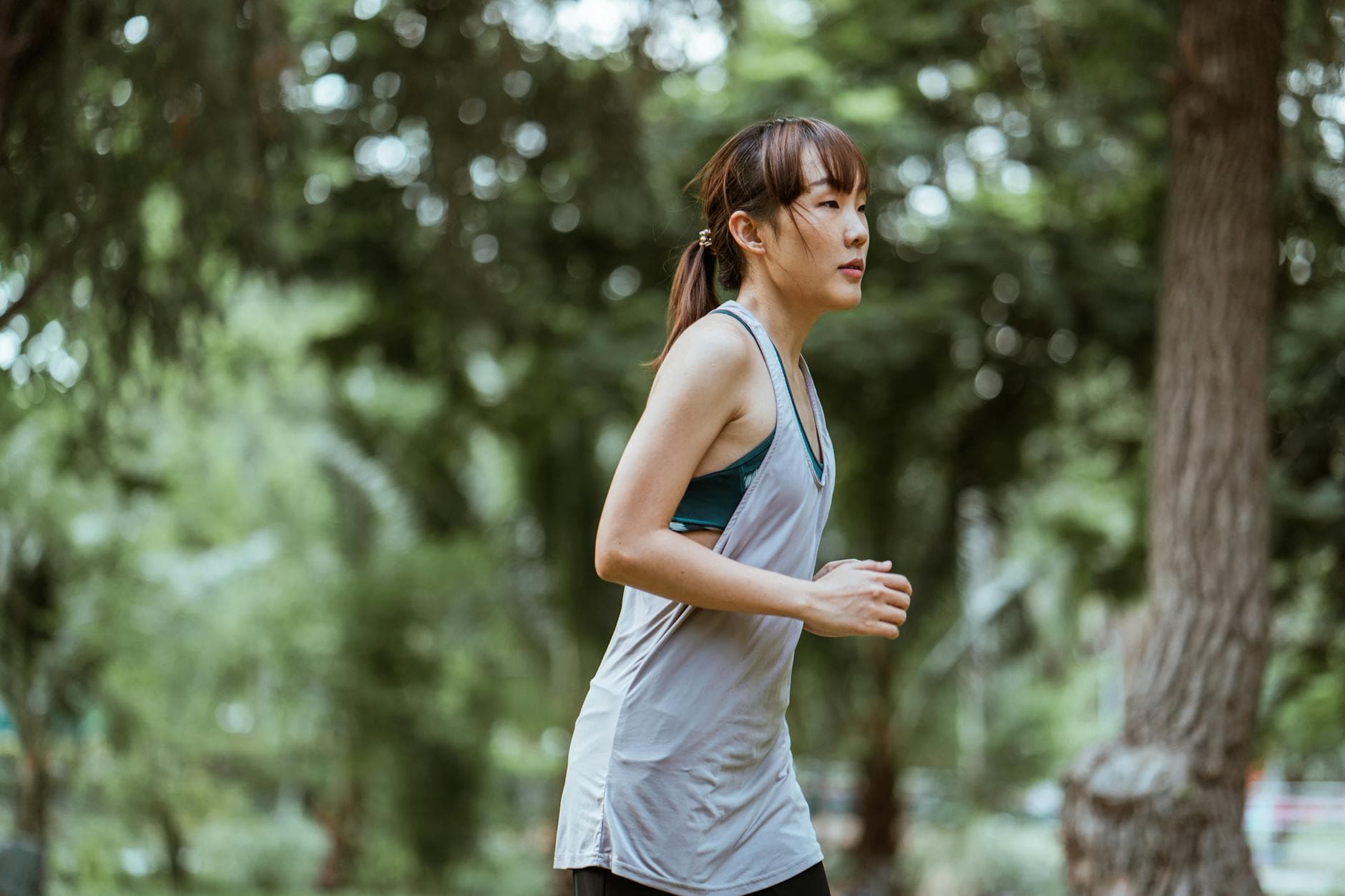 Side view of determined young Asian sportswoman in activewear running in green summer park on clear day and looking away thoughtfully - spring cardio fat burn