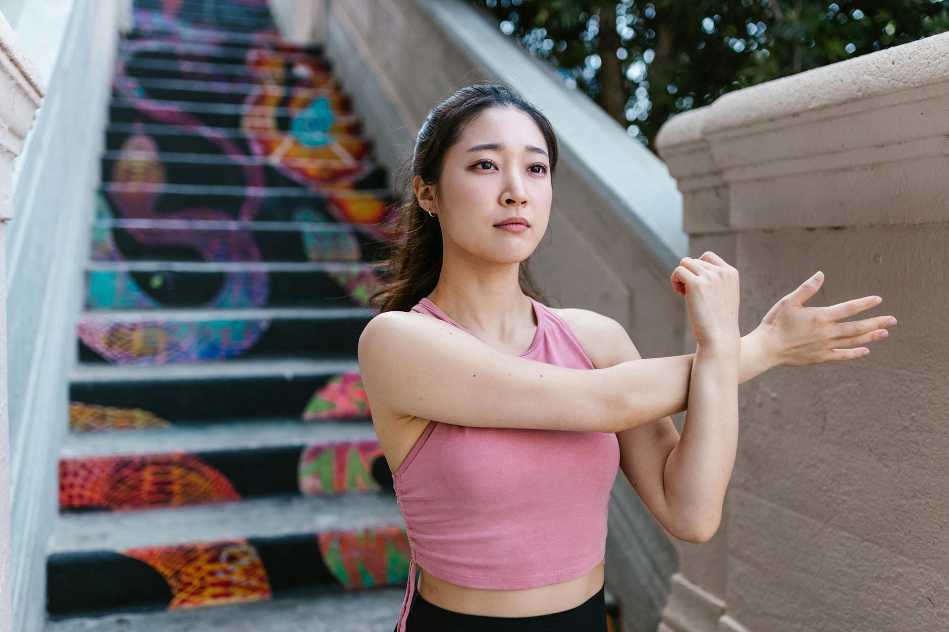 A focused young woman in a pink crop top stretches on colorful graffiti stairs. - desk stretches
