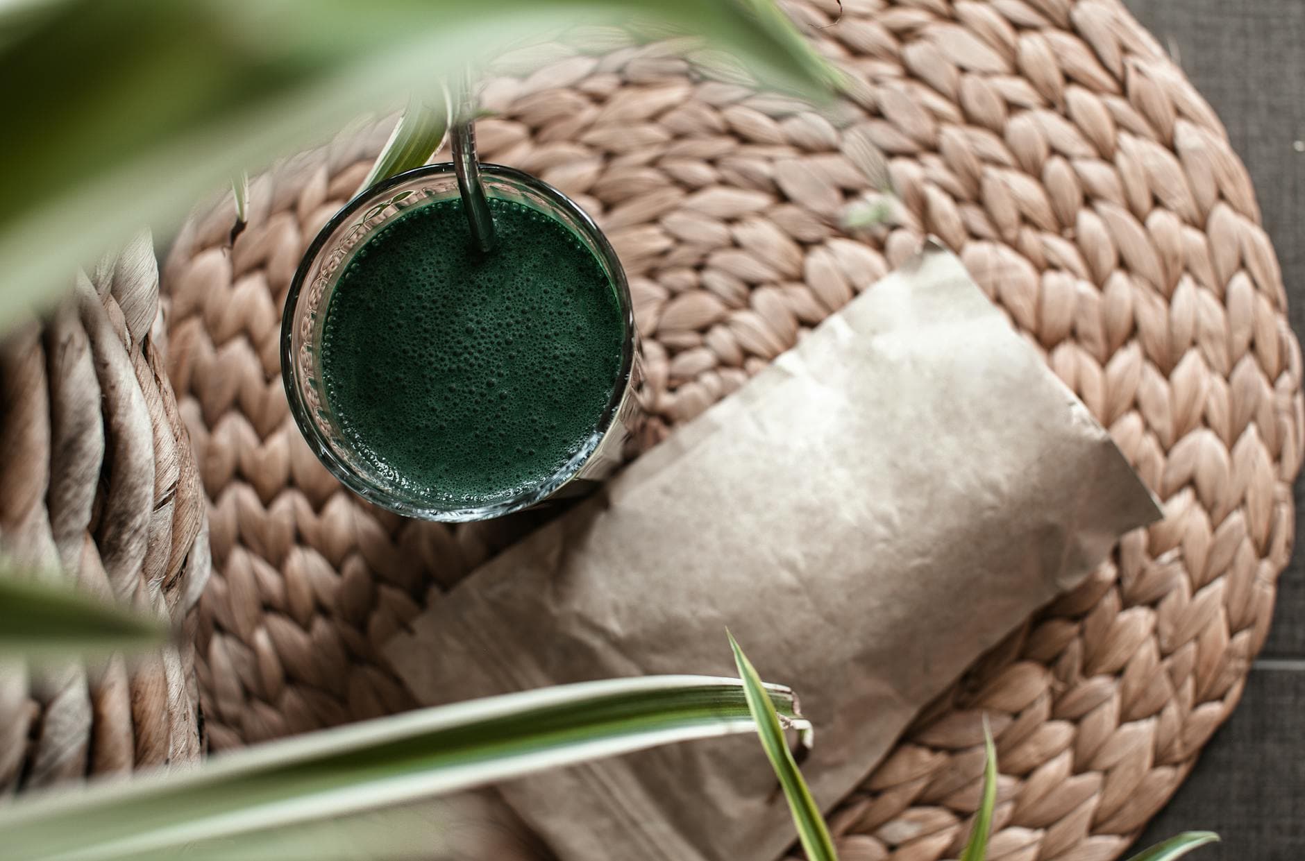 Aerial view of a frothy green matcha drink in a glass on a woven mat, surrounded by green leaves. - green smoothie benefits