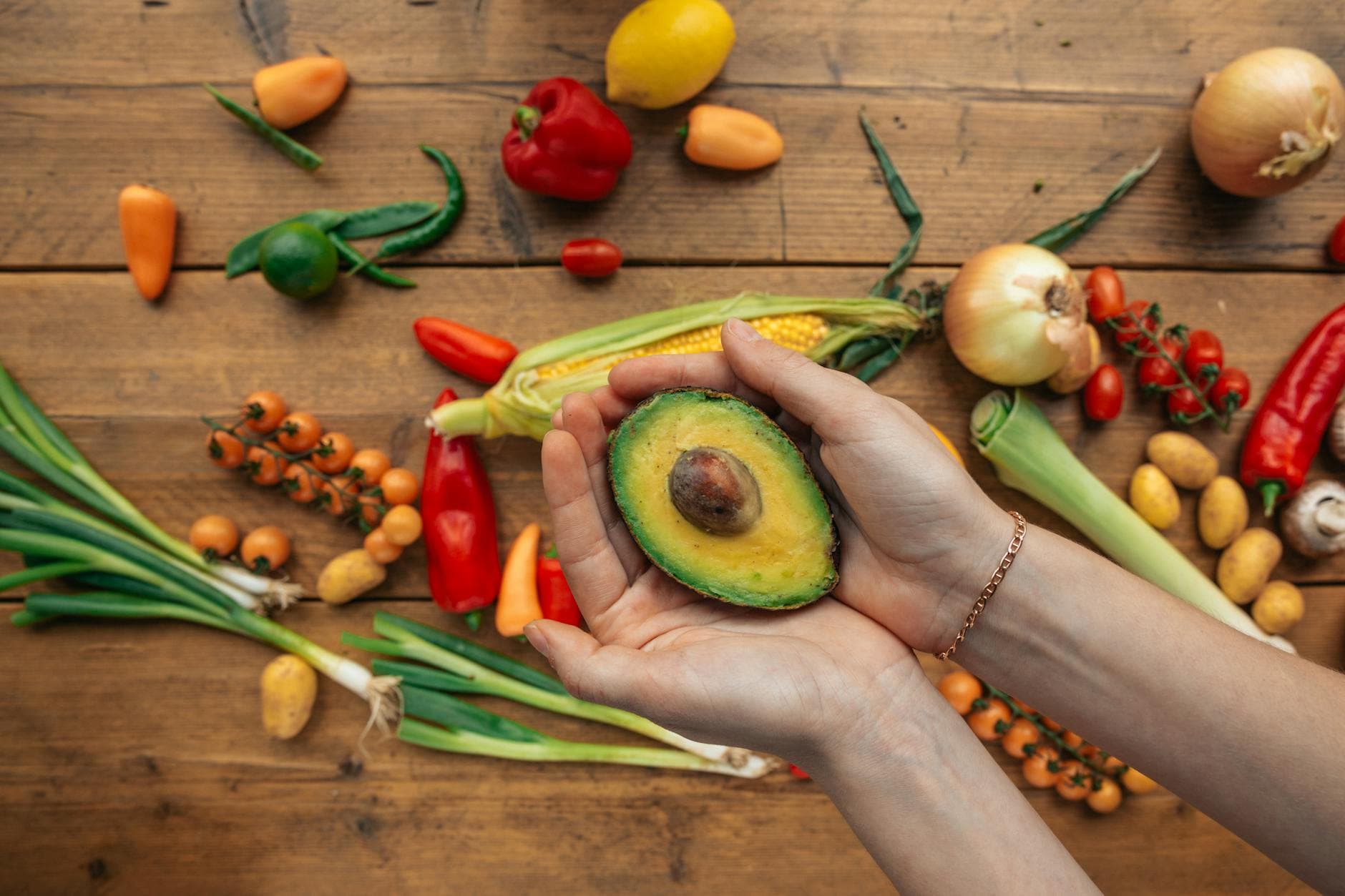 Sliced avocado held in hands surrounded by vibrant fresh vegetables on wooden background. - macronutrient balance spring