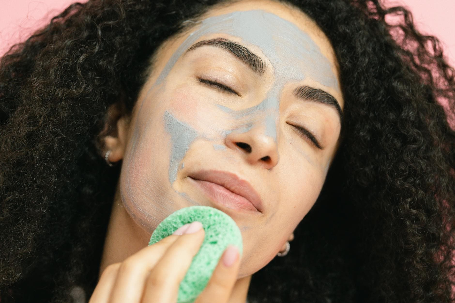 A woman with curly hair enjoys a relaxing facial treatment with a clay mask and sponge. - spring skincare tips