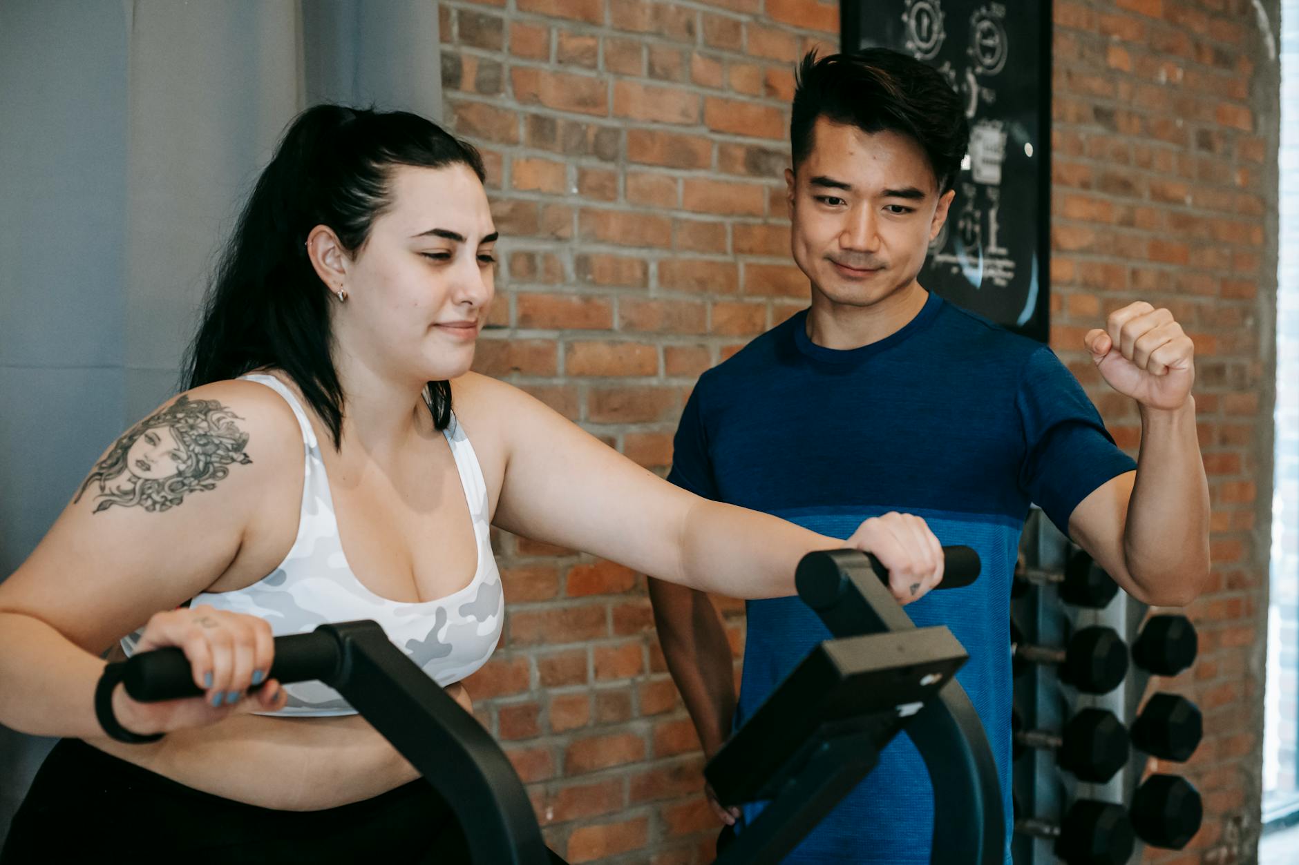 Smiling young tattooed plus size sportswoman in activewear doing cardio exercise on fitness cycle while training in contemporary gym with positive Asian male instructor - best cardio for weight loss