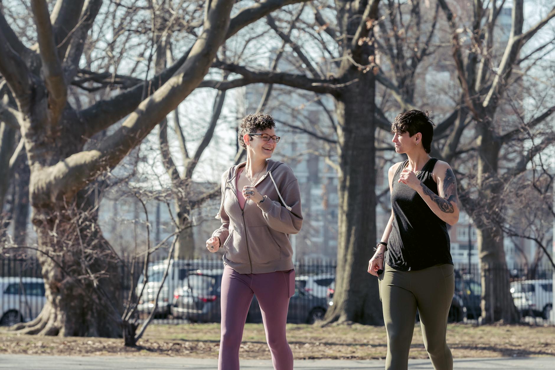 Glad sporty women smiling and looking at each other while running in park of town with leafless trees - best morning exercises