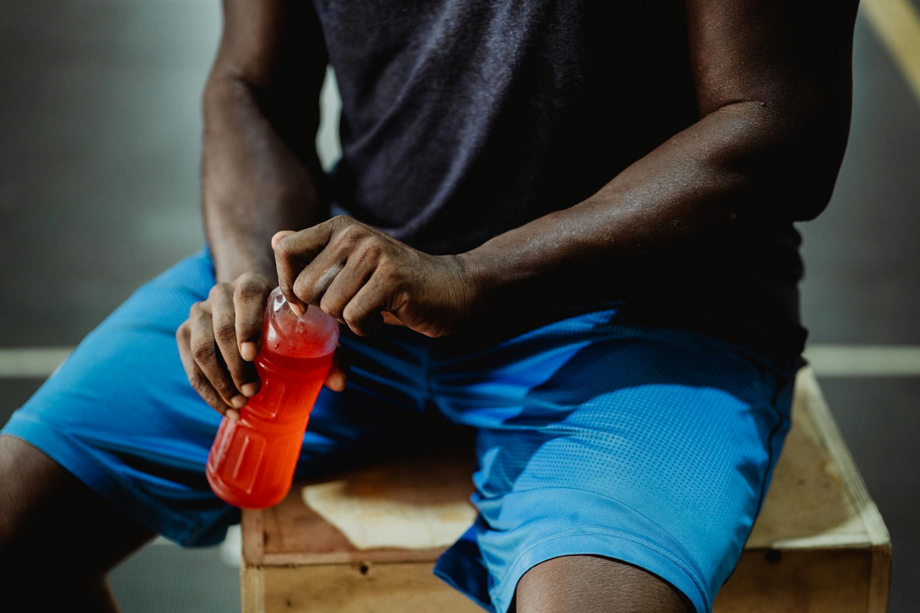 A man resting on a wooden box, enjoying a red sports drink during his workout session. - best post workout drink
