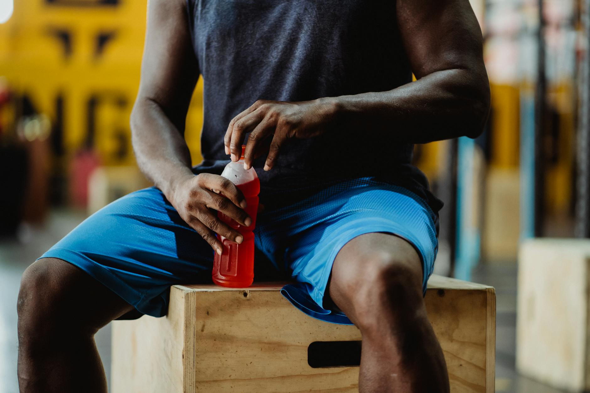 A man sitting on a wooden box, holding a red sports drink, taking a break from exercise. - best post workout drink