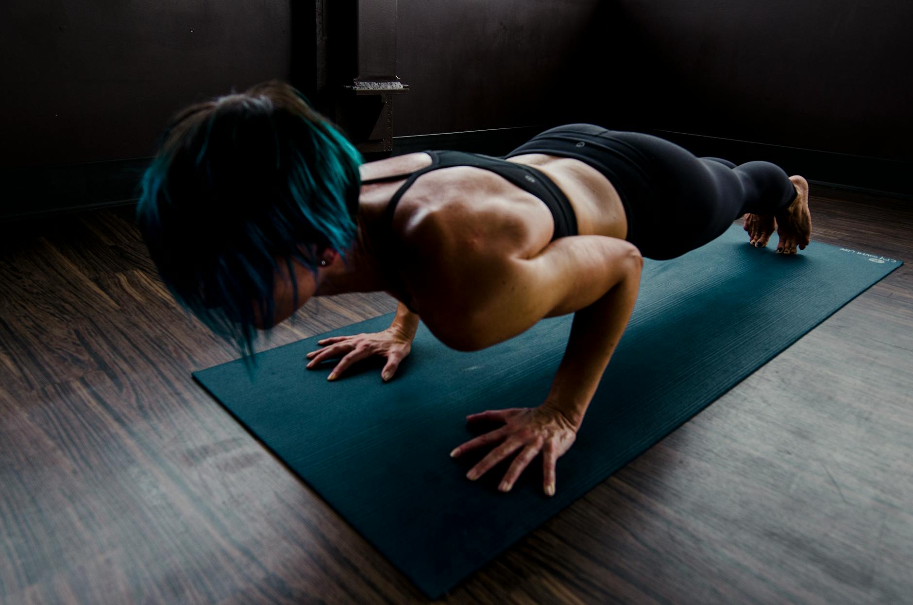 A fit woman with blue hair performs a push-up on a yoga mat indoors, showcasing strength and focus. - core workout at home