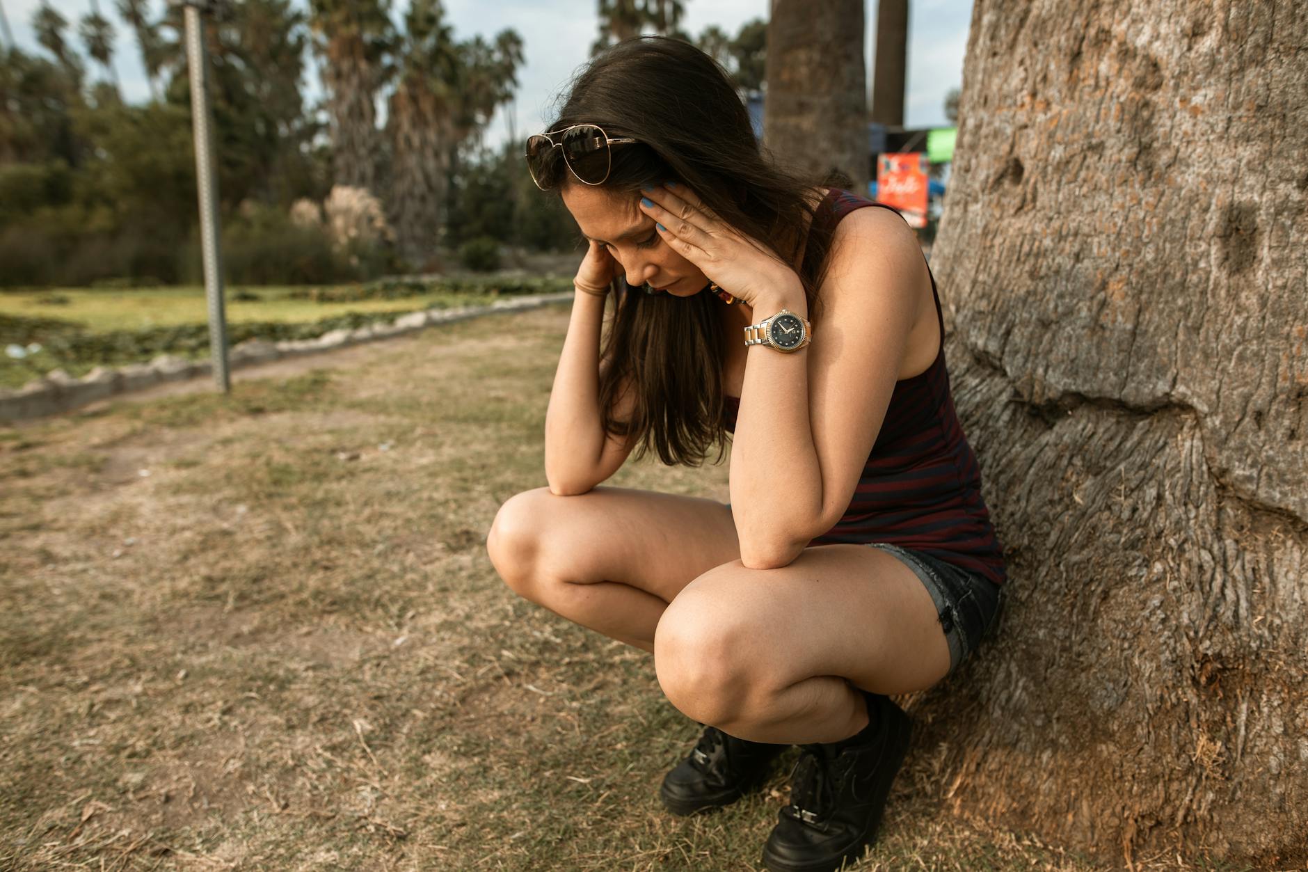 A woman kneels by a tree holding her head, expressing stress or anxiety in an outdoor park. - deep breathing for anxiety