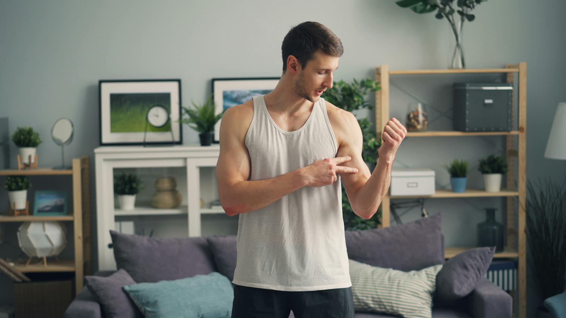 Muscular man in a tank top standing indoors, examining his biceps with a modern living room background. - flexibility exercise examples