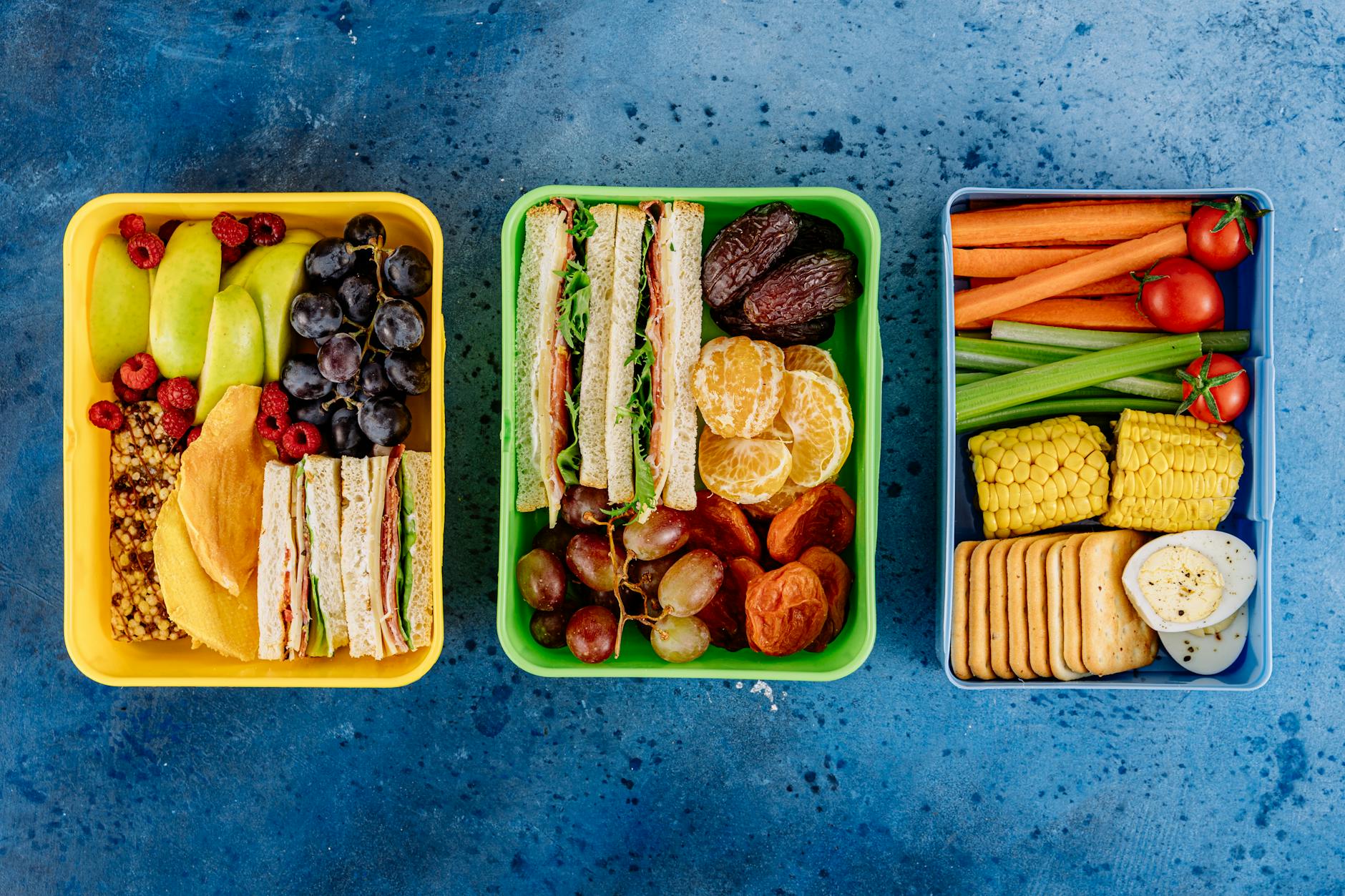 Three vibrant lunch boxes showcasing fruits, sandwiches, and vegetables on a blue textured background. - healthy meal plan ideas