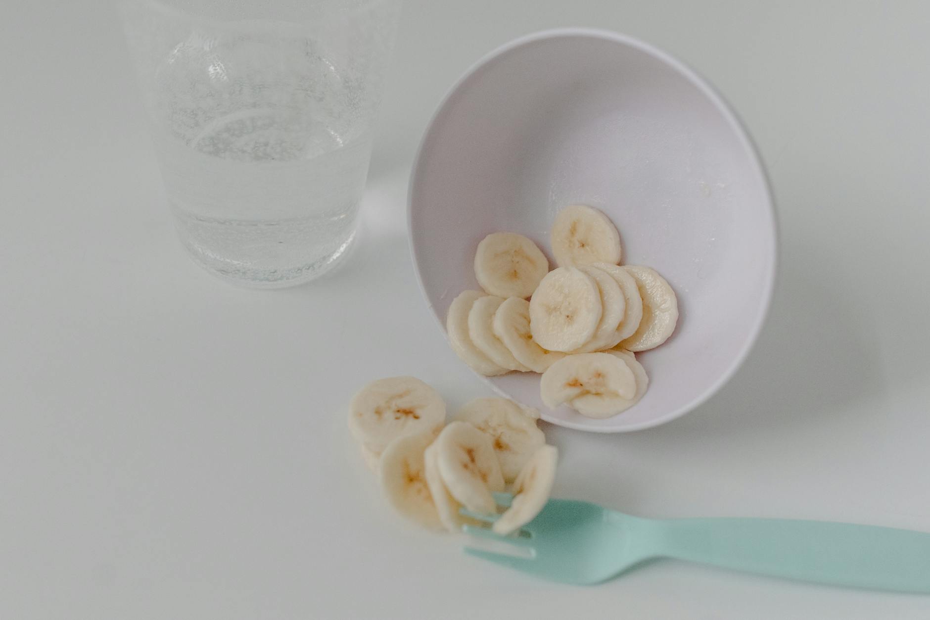 Sliced banana pieces in a white bowl with a mint fork and glass of water on a clean table. - healthy snack ideas