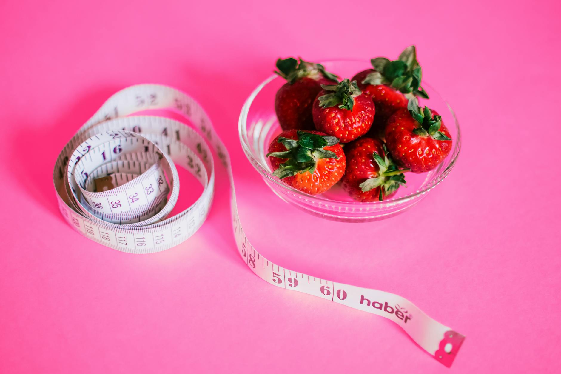 A glass bowl of fresh strawberries paired with a pink measuring tape symbolizes healthy eating. - healthy snacks for weight loss