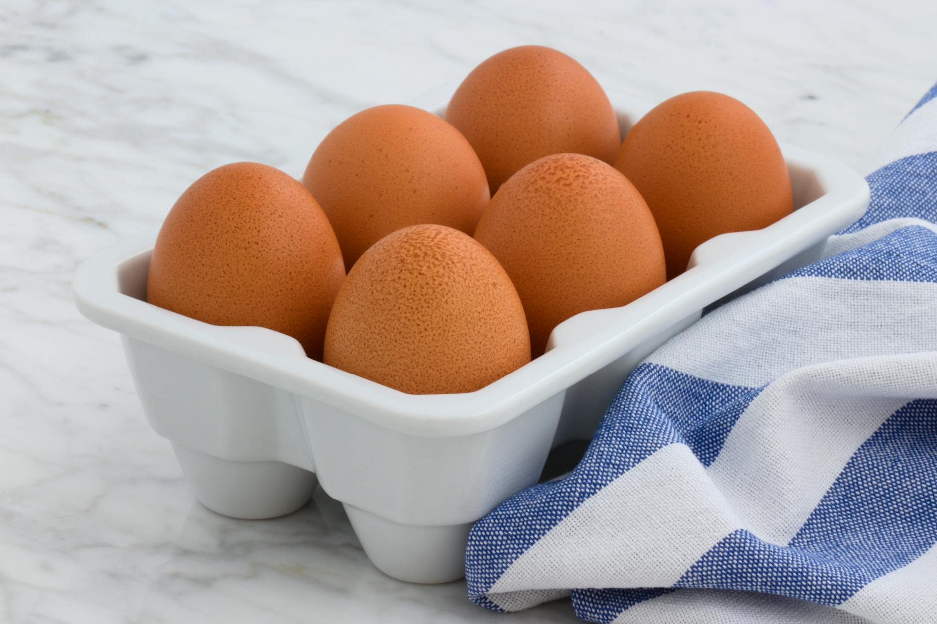 A close-up of fresh brown eggs arranged in a ceramic tray with a striped cloth on a marble surface. - high protein foods