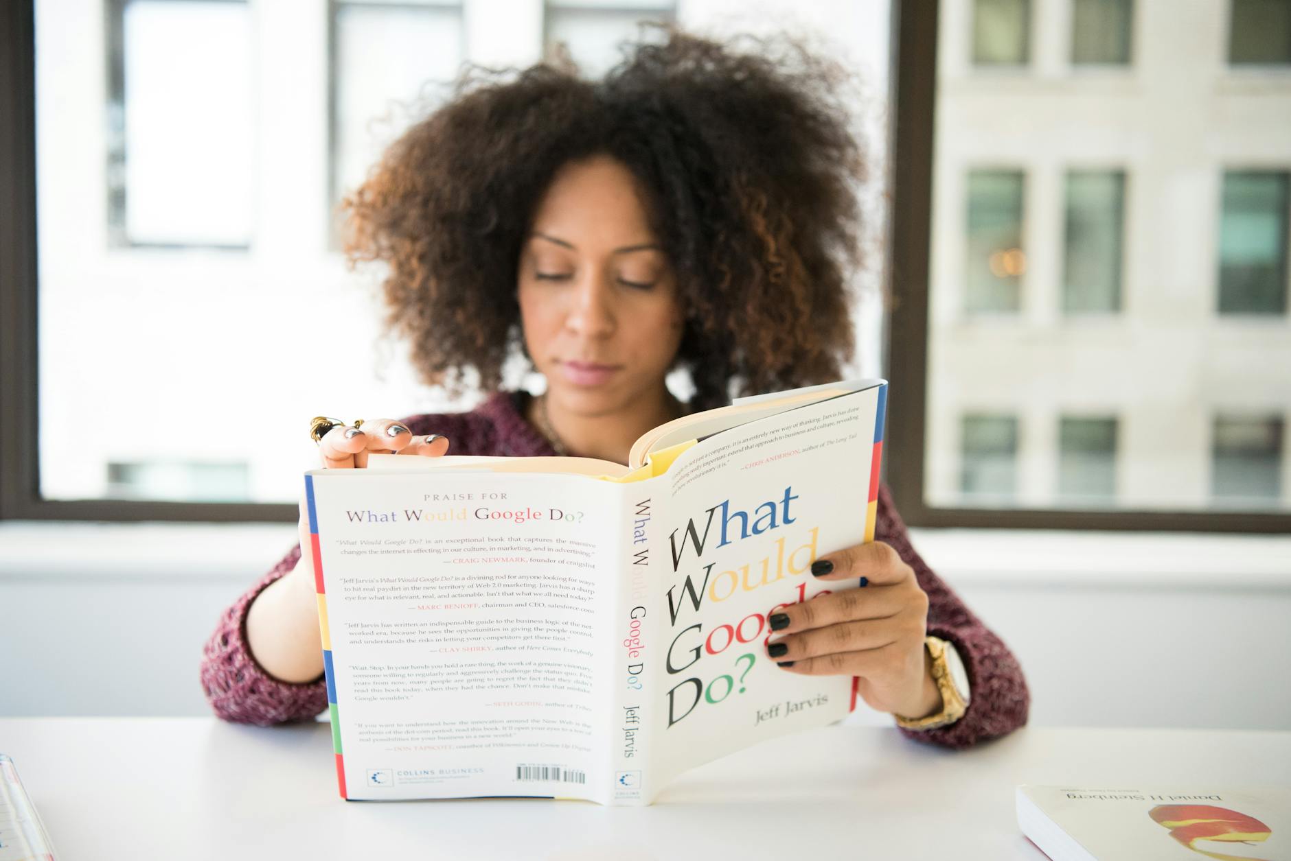 A woman reading 'What Would Google Do?' at a desk by a window in a modern office. - what is meditation