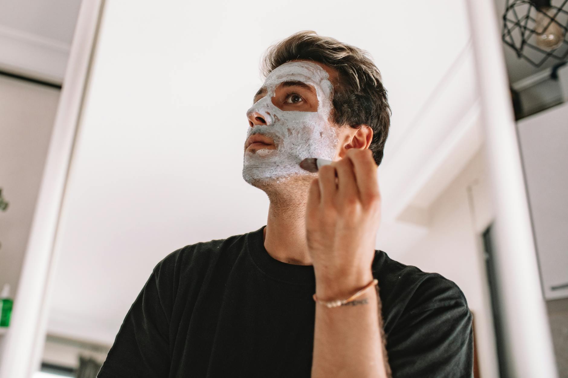 Adult man applying a facial mask while looking in the mirror at home, promoting skincare routine. - men's natural skincare