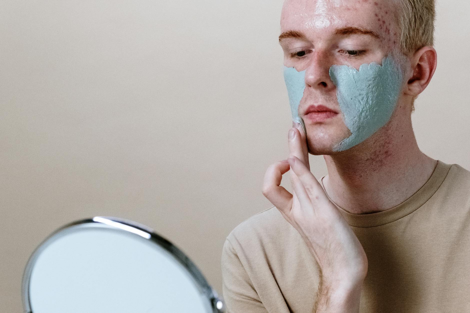 Close-up of a young man applying a clay face mask with a mirror. - natural acne treatment