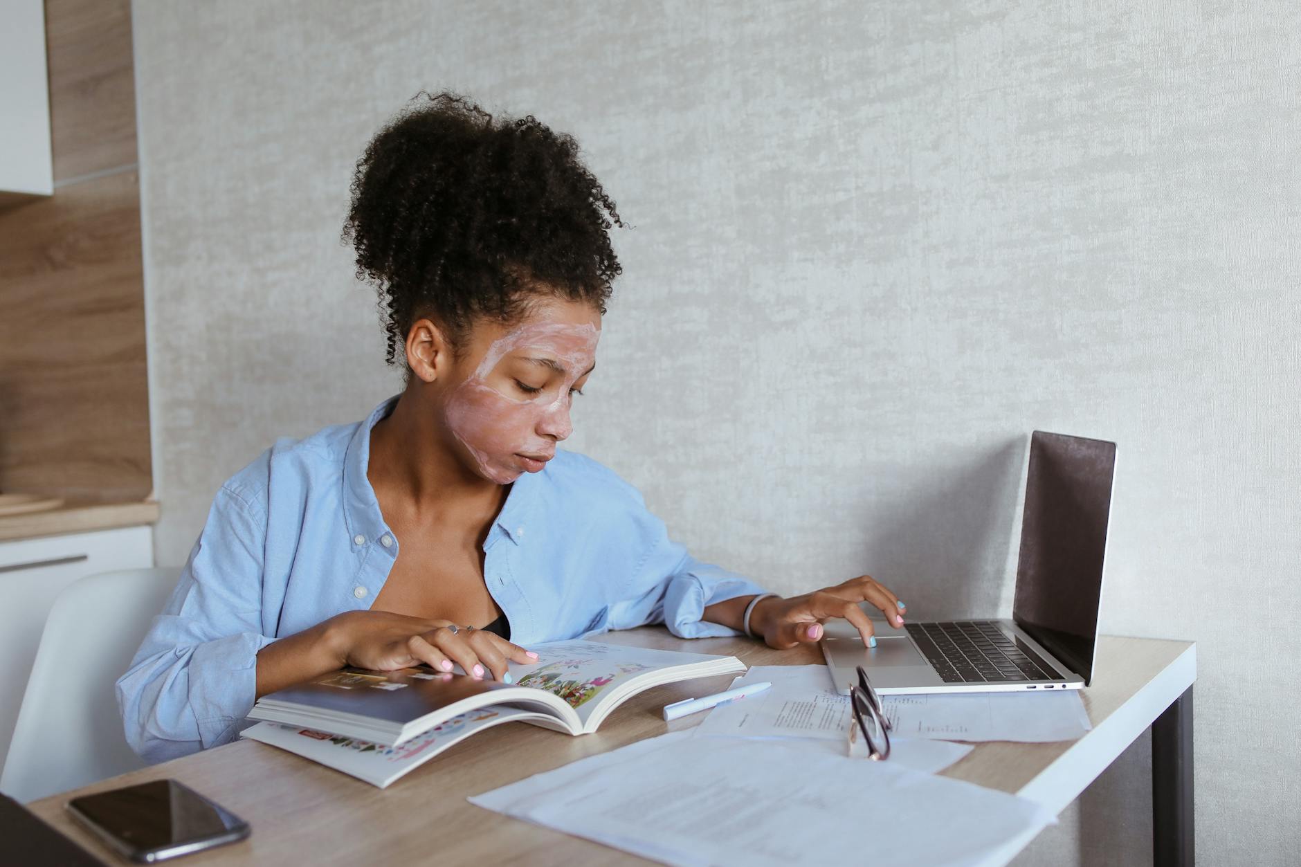 A woman multitasks with a laptop and book while applying skincare at home. - just natural skincare reviews