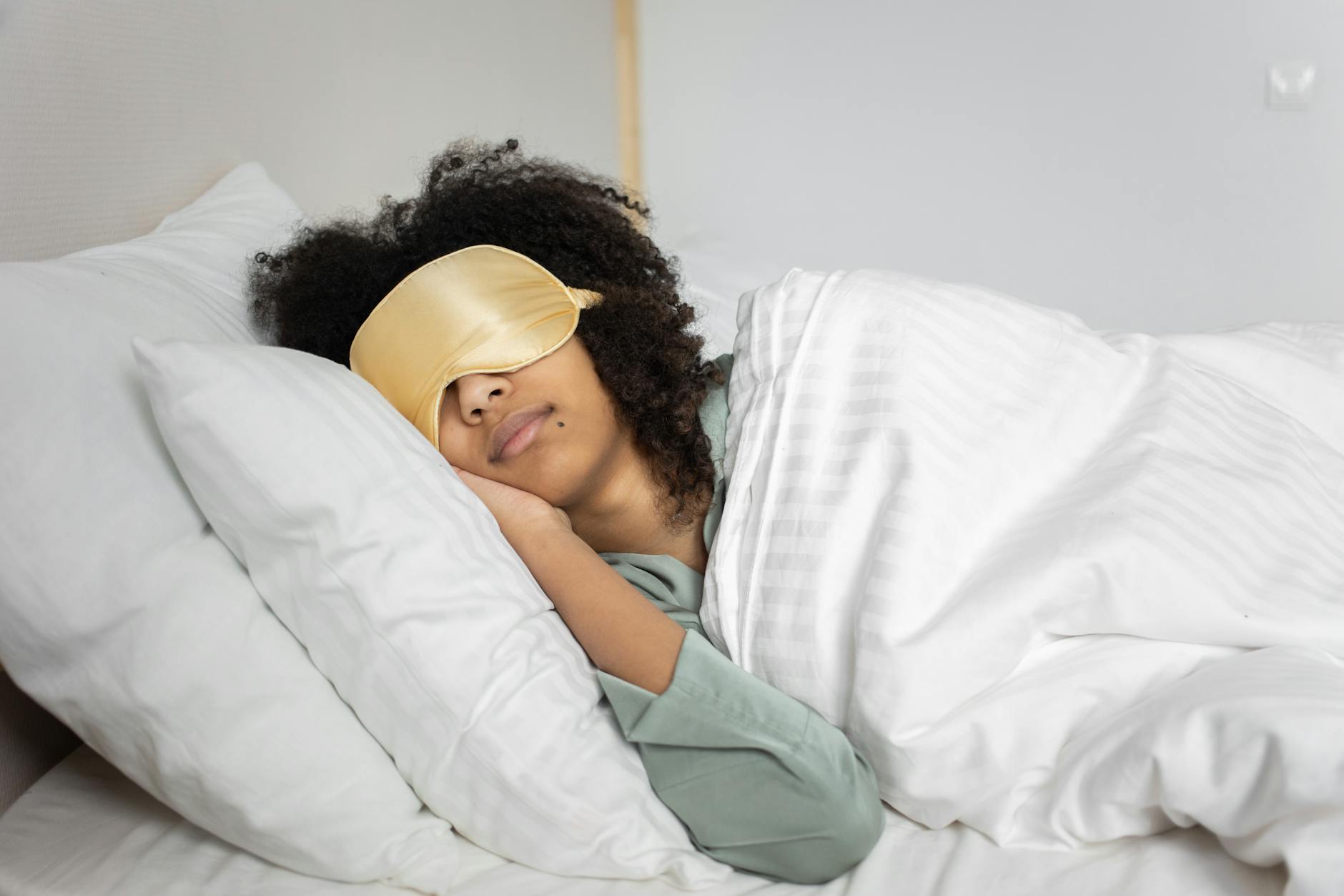 A woman with afro hair sleeps soundly in bed with a sleep mask, enjoying a cozy indoor atmosphere. - what is sleep hygiene