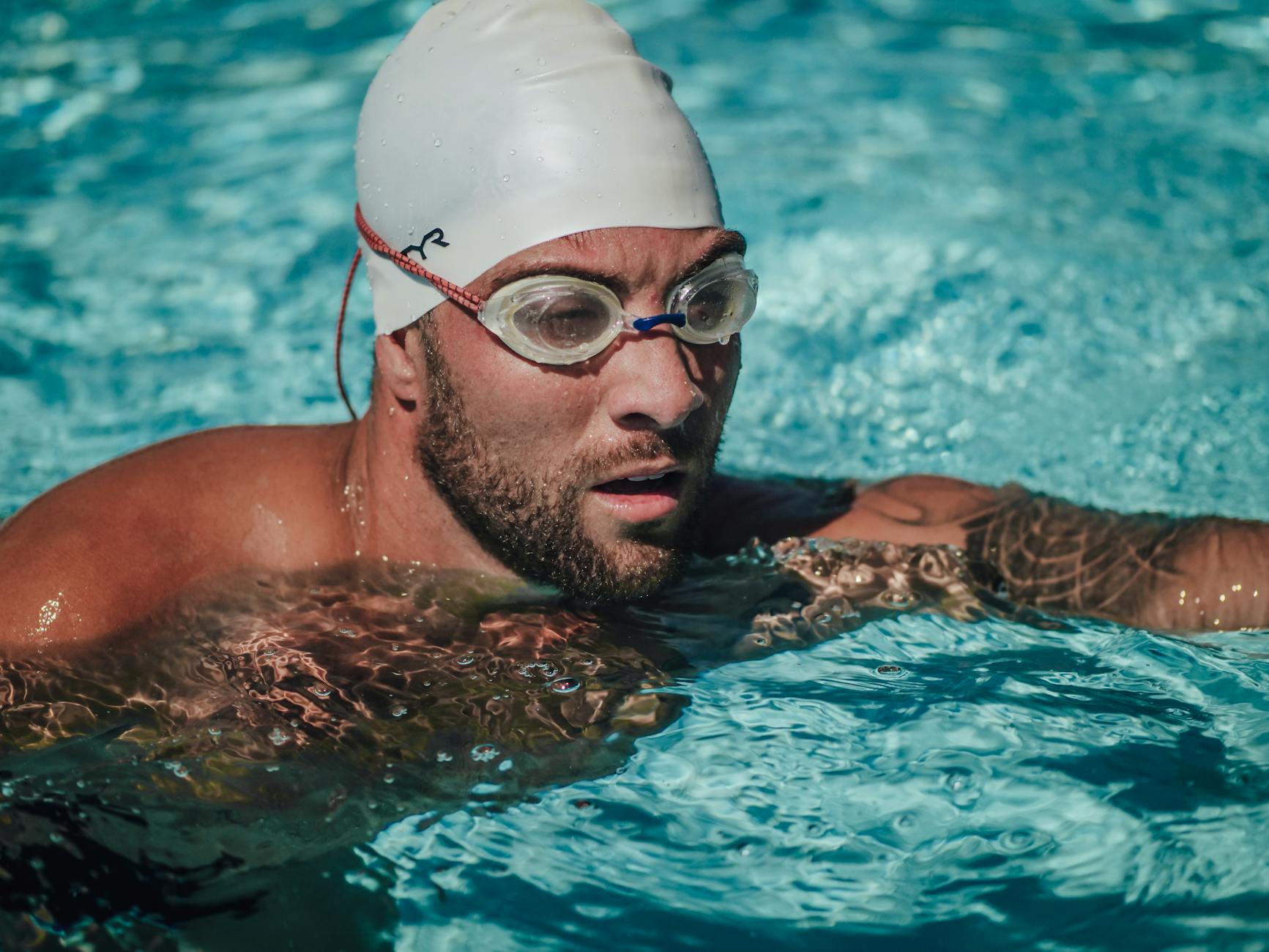 A focused male swimmer with tattoos and beard wearing goggles and a swimming cap in a clear blue pool. - swimming benefits for men