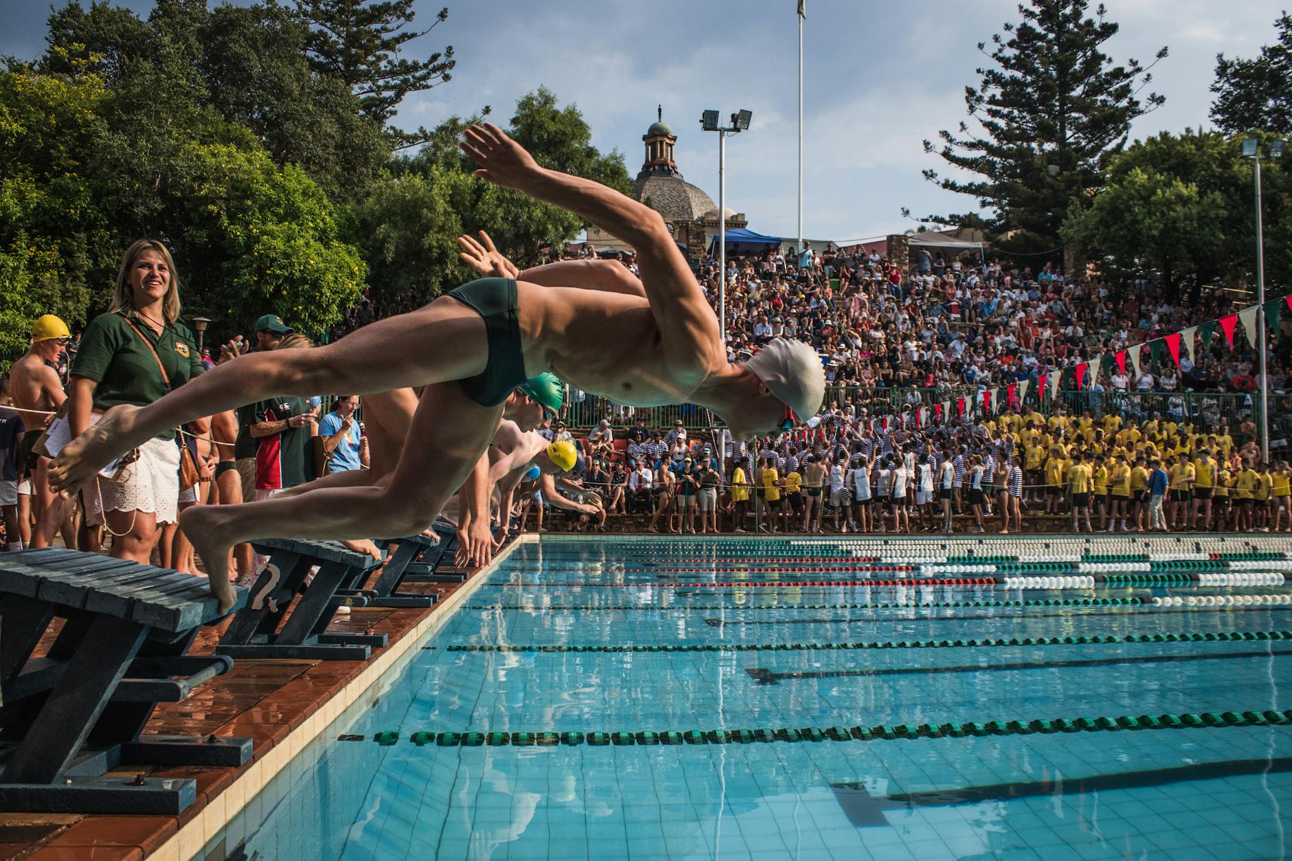 Swimmers diving into an outdoor pool during a competitive sporting event in front of a large crowd. - swimming benefits for men
