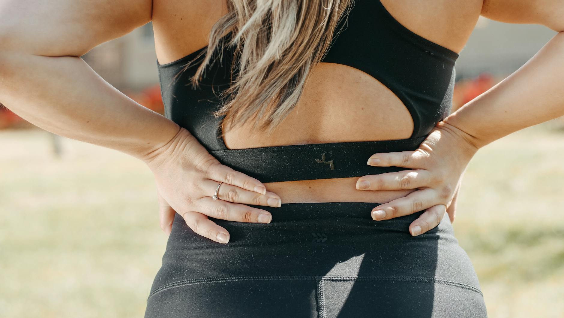 A woman in activewear holds her lower back, indicating muscle strain, outdoors. - back pain stretches