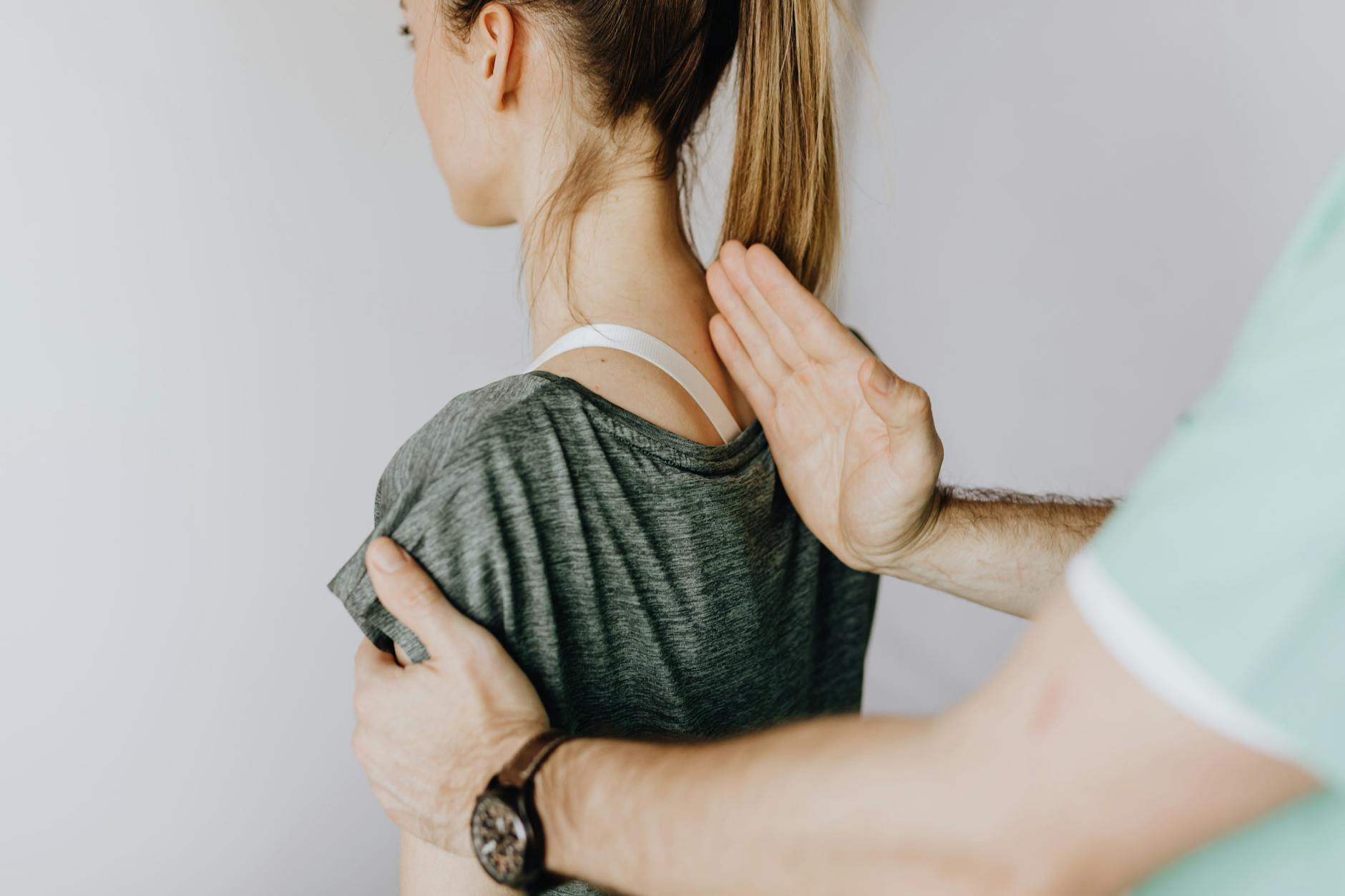 Back view of crop unrecognizable osteopath in uniform and wristwatch checking up back of slim female patient in casual wear on white background - bad posture correction
