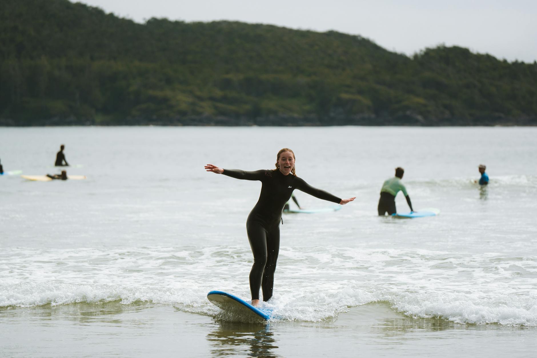 An excited woman surfing on a calm beach with others in view, showcasing outdoor fun. - beginner exercises