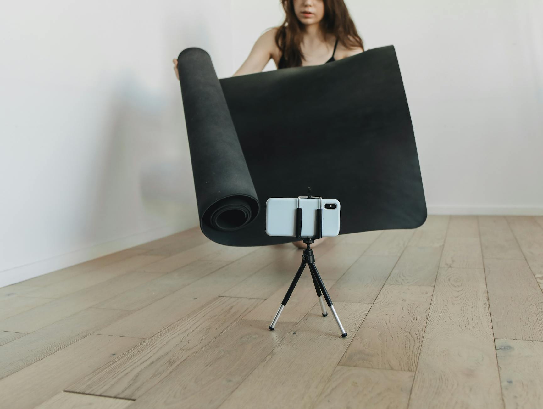 Woman rolls out yoga mat in living room with smartphone tripod for online session. - beginner yoga video