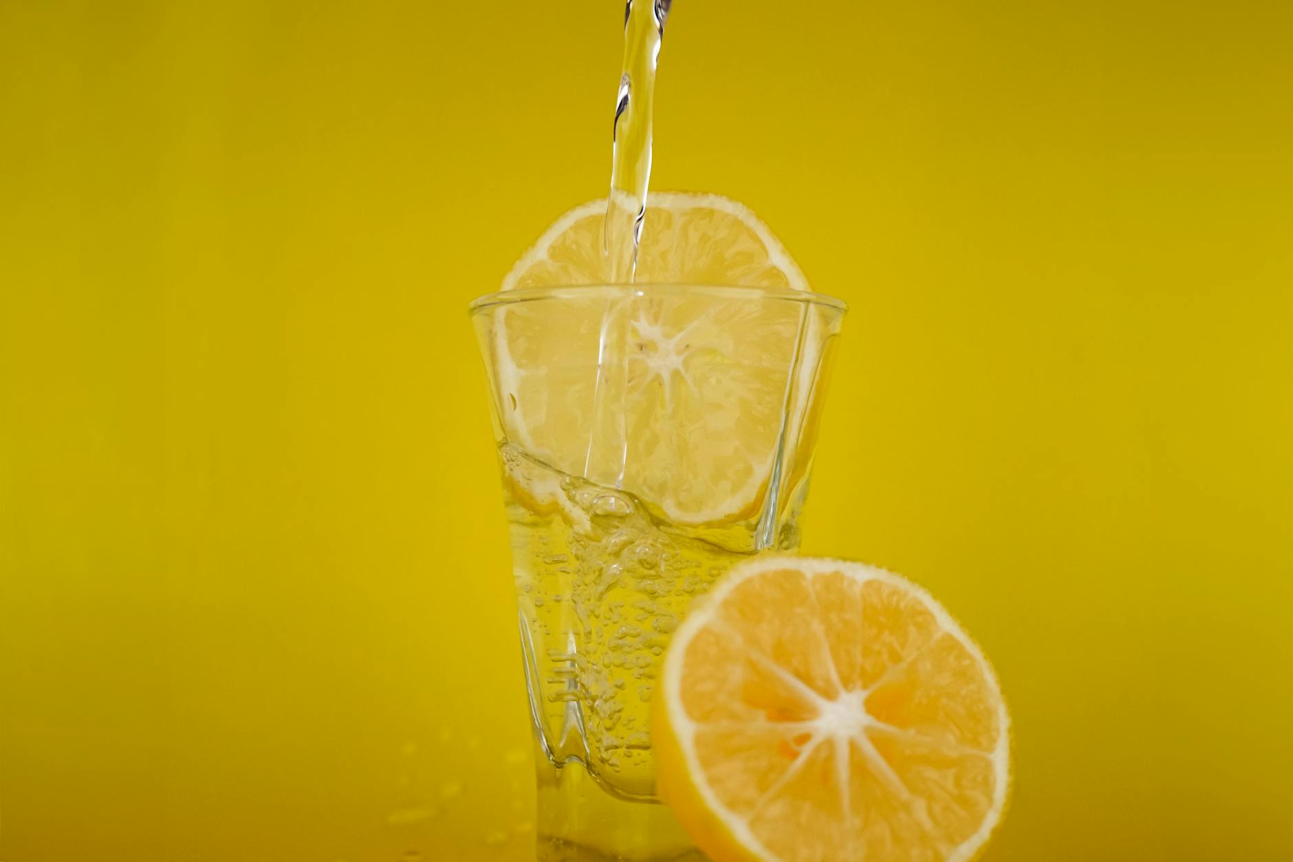 A close-up of water pouring into a glass with lemon slices, vibrant yellow backdrop. - best detox water