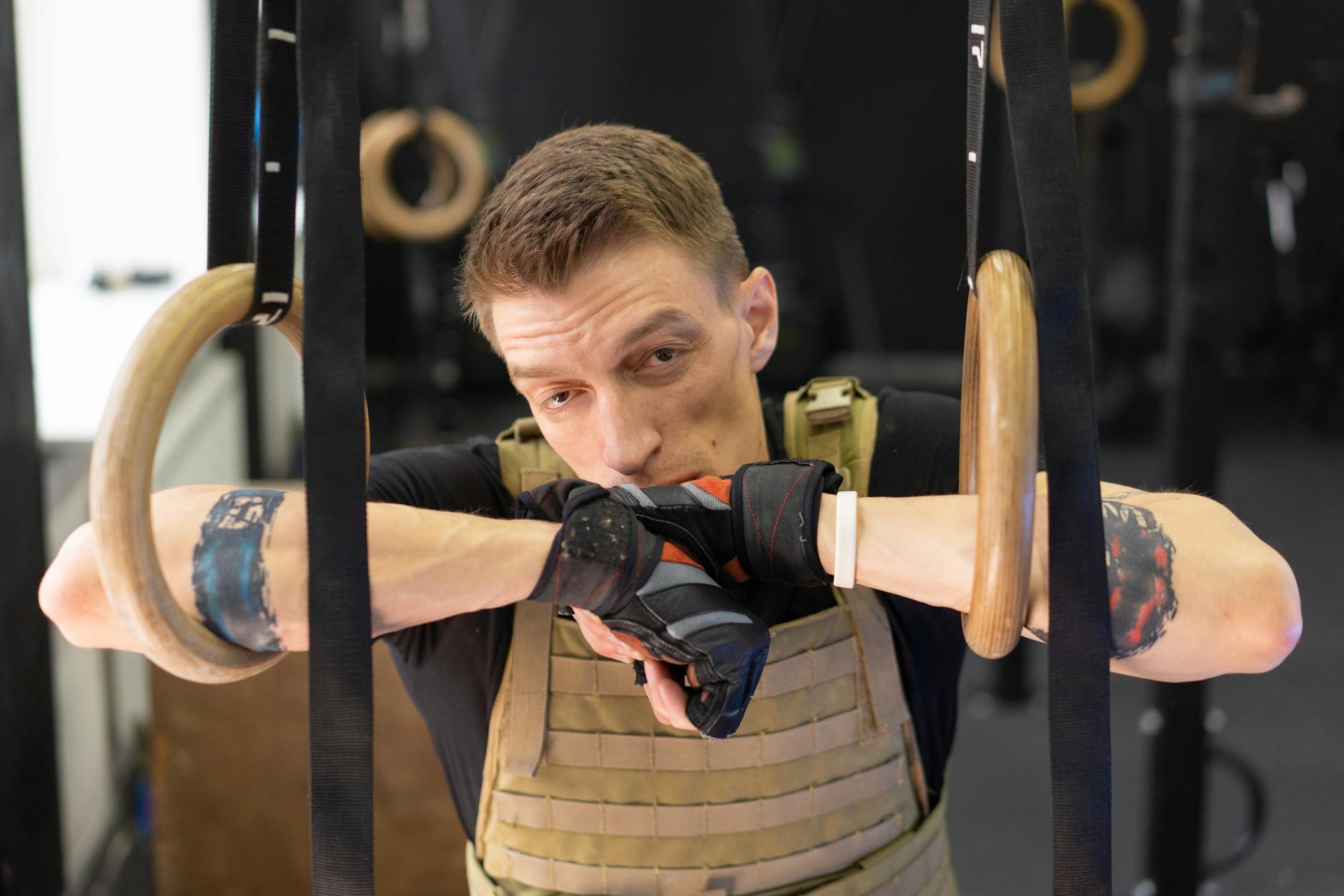 Adult male athlete taking a break on gymnastic rings wearing tactical vest and workout gloves indoors. - bodyweight arm exercises