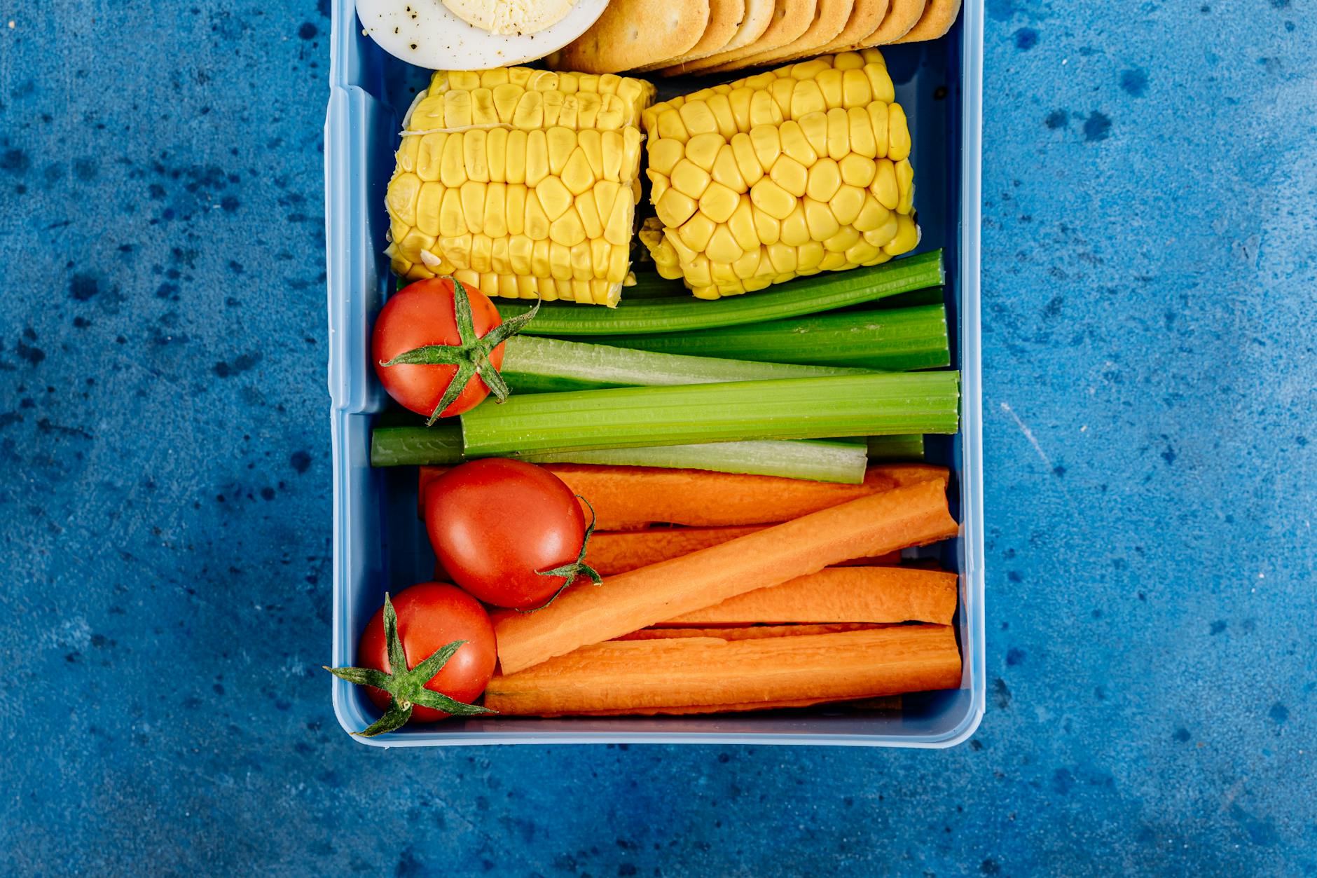 Top view of a lunchbox filled with vegetables, corn, tomatoes, and snacks on a blue background. - easy healthy meals