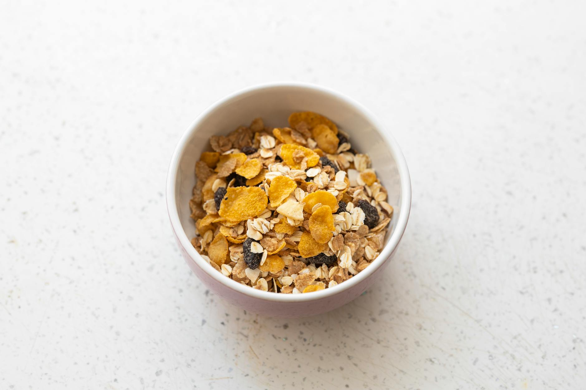 A ceramic bowl filled with mixed cereals including cornflakes, oats, and raisins on a white surface. - easy healthy meals