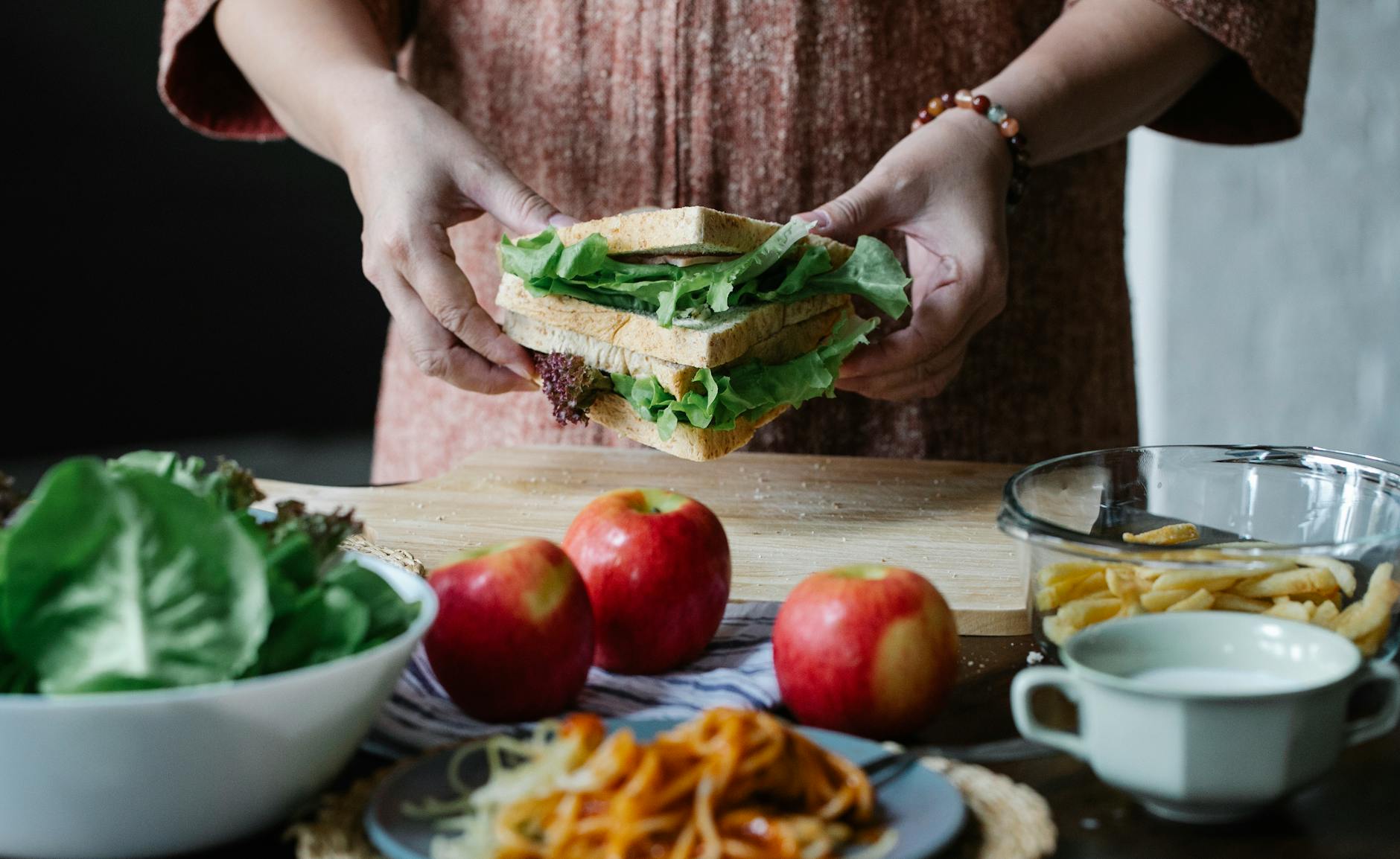 Unrecognizable female standing at table with apples and cutting board with sandwich with lettuce in hands in kitchen on breakfast time - easy healthy recipes