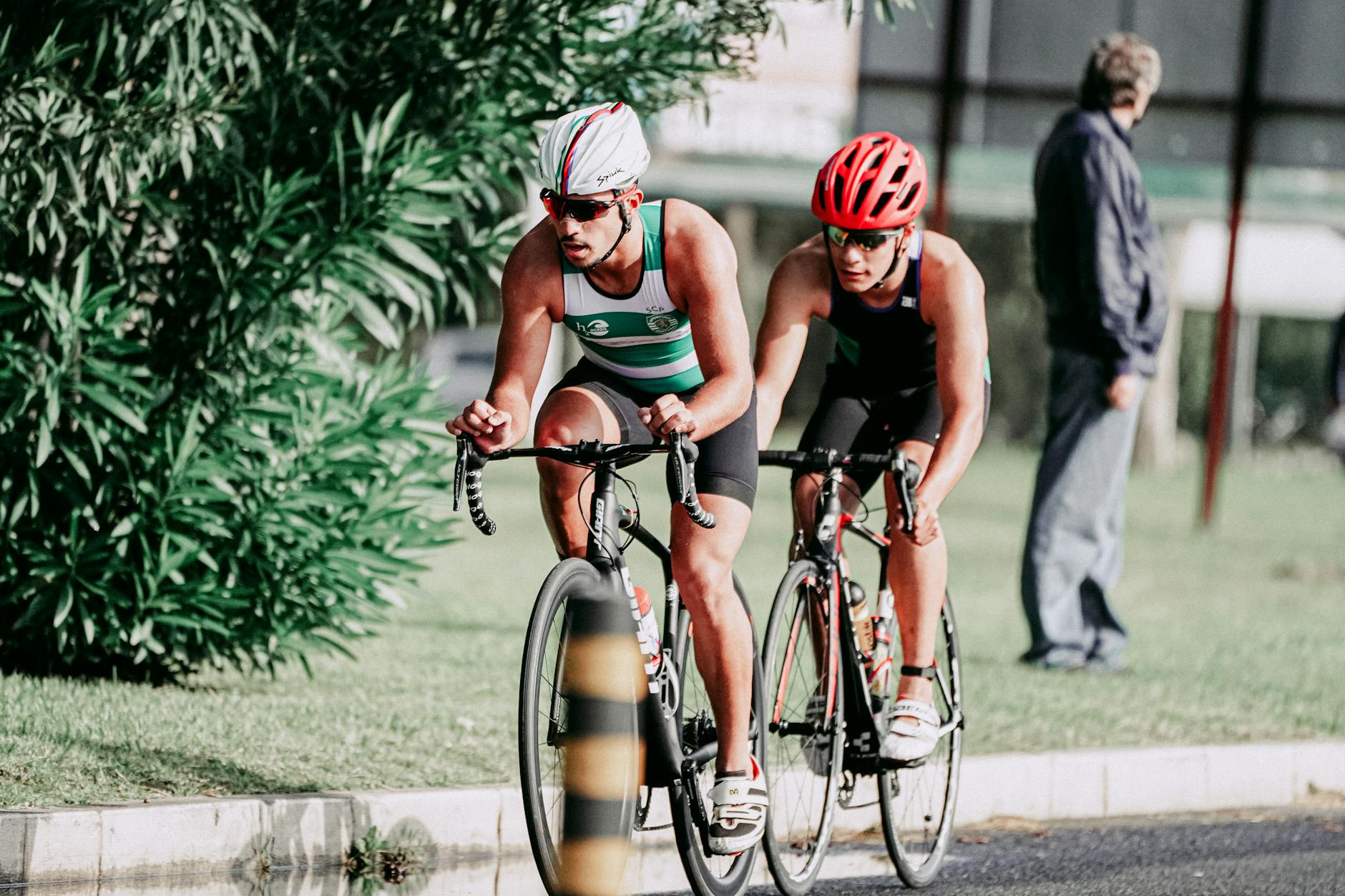 Full body of concentrated tanned bikers in comfortable t shirts and shorts riding on bicycles on asphalt road along vibrant green tree in sunlight - endurance cycling training