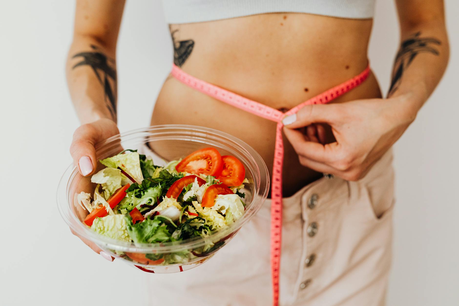 Woman holding vegetable salad and measuring waist with tape, symbolizing healthy lifestyle choices. - fat burning foods