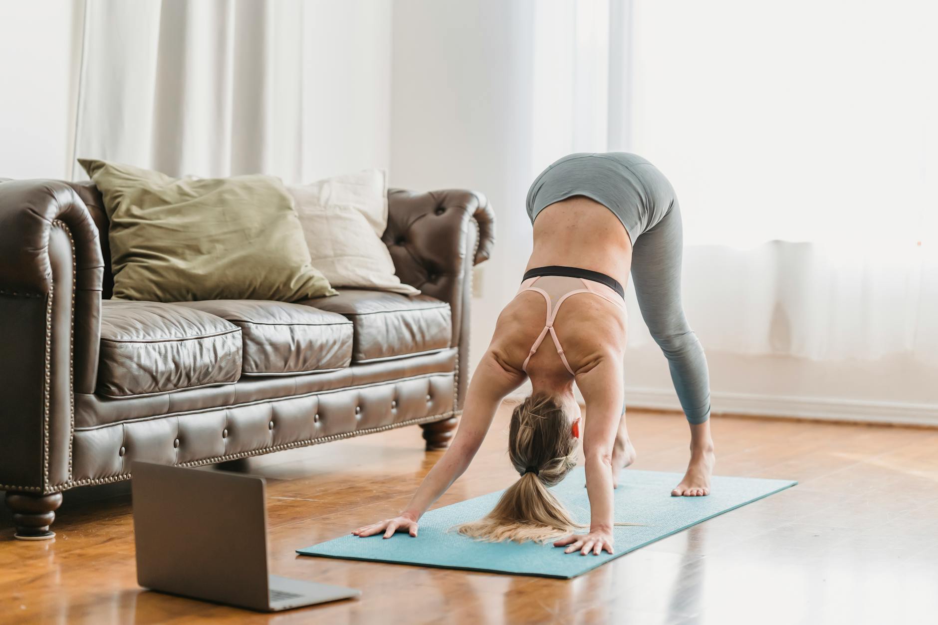Full length of faceless lady wearing leggings and sportive bra stretching back on yoga mat near opened netbook and leather couch with pillows in light living room near curtains - flexibility exercises list