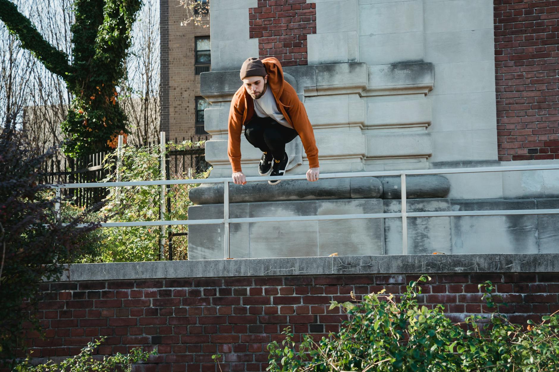 Full body determined young male tracer wearing hoodie and hat jumping over metal railing near old break building in sunlight - free running beginners