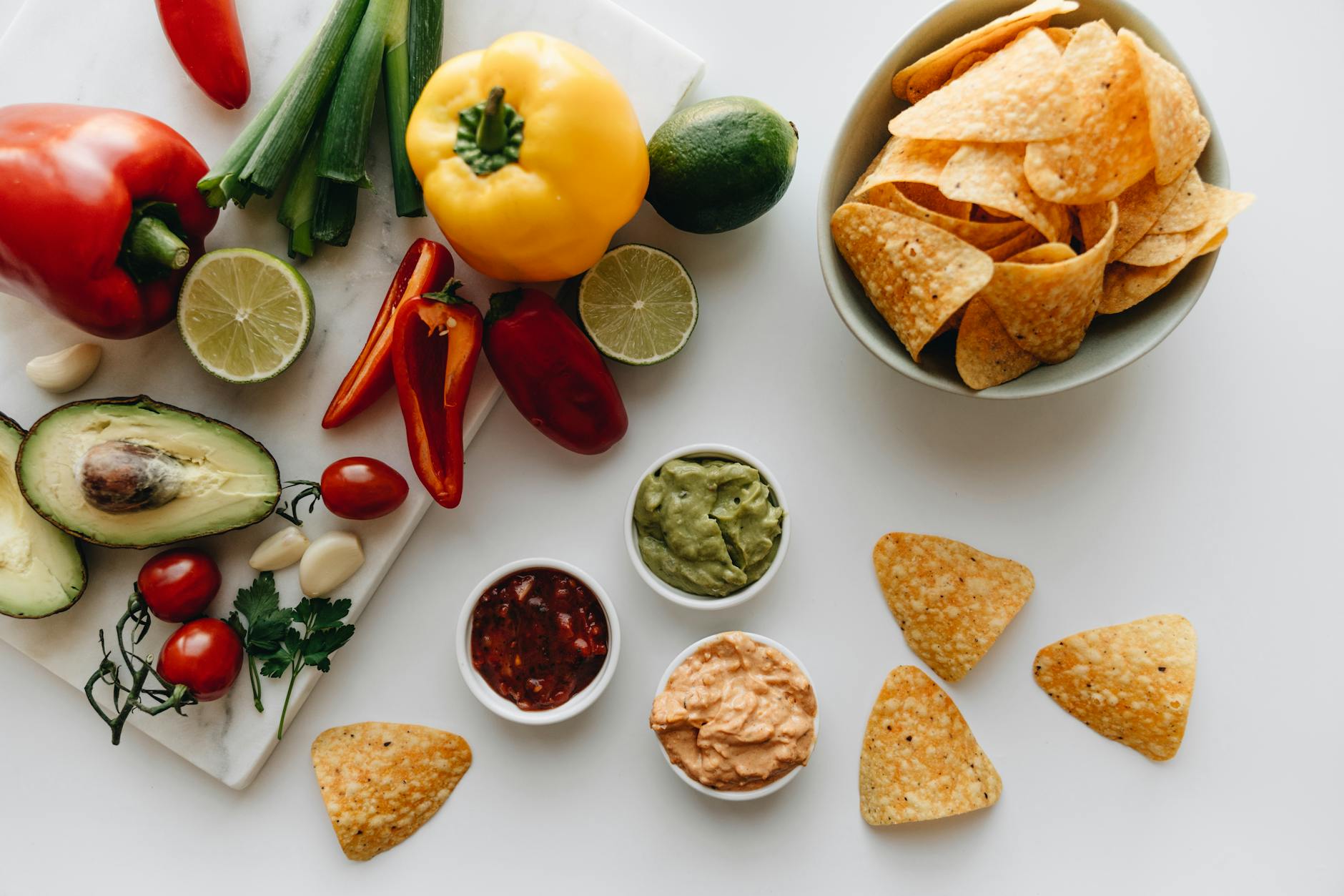 Colorful assortment of fresh vegetables and snacks displayed on a white background with dips. - healthy chips