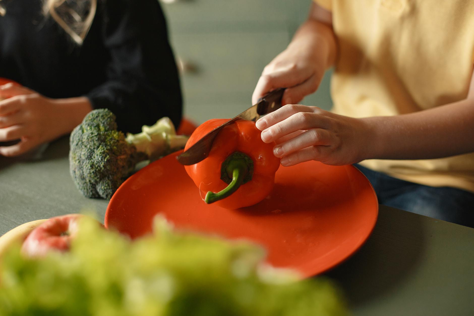 Unrecognizable boy and girl cutting red pepper on plate sitting at table with various vegetables and fruits while preparing healthy food - healthy kid meals