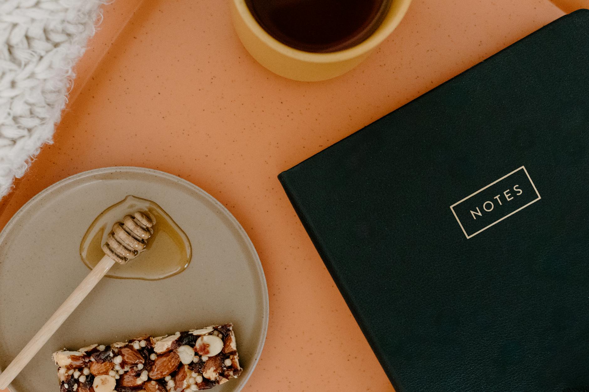 Top view of a cozy setup with a notebook, mug, honey, and snack on a warm orange surface. - healthy office snacks
