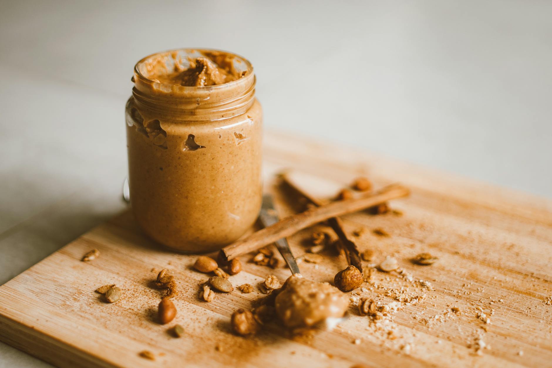 A jar of peanut butter with scattered peanuts and cinnamon on a cutting board. - healthy peanut butter snacks