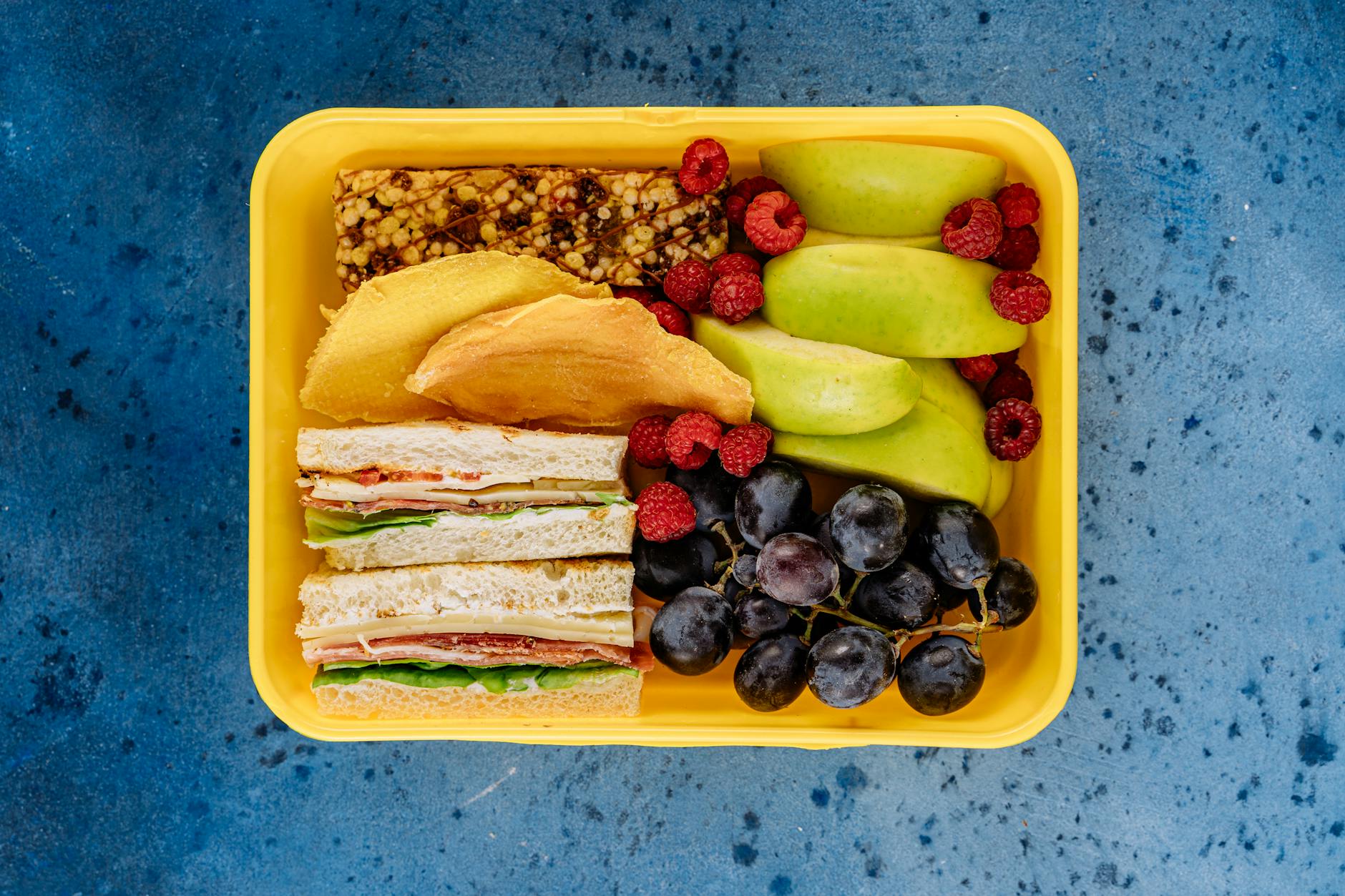 Colorful lunch box with fruits, sandwiches, and a snack bar on a blue background. - healthy school lunches