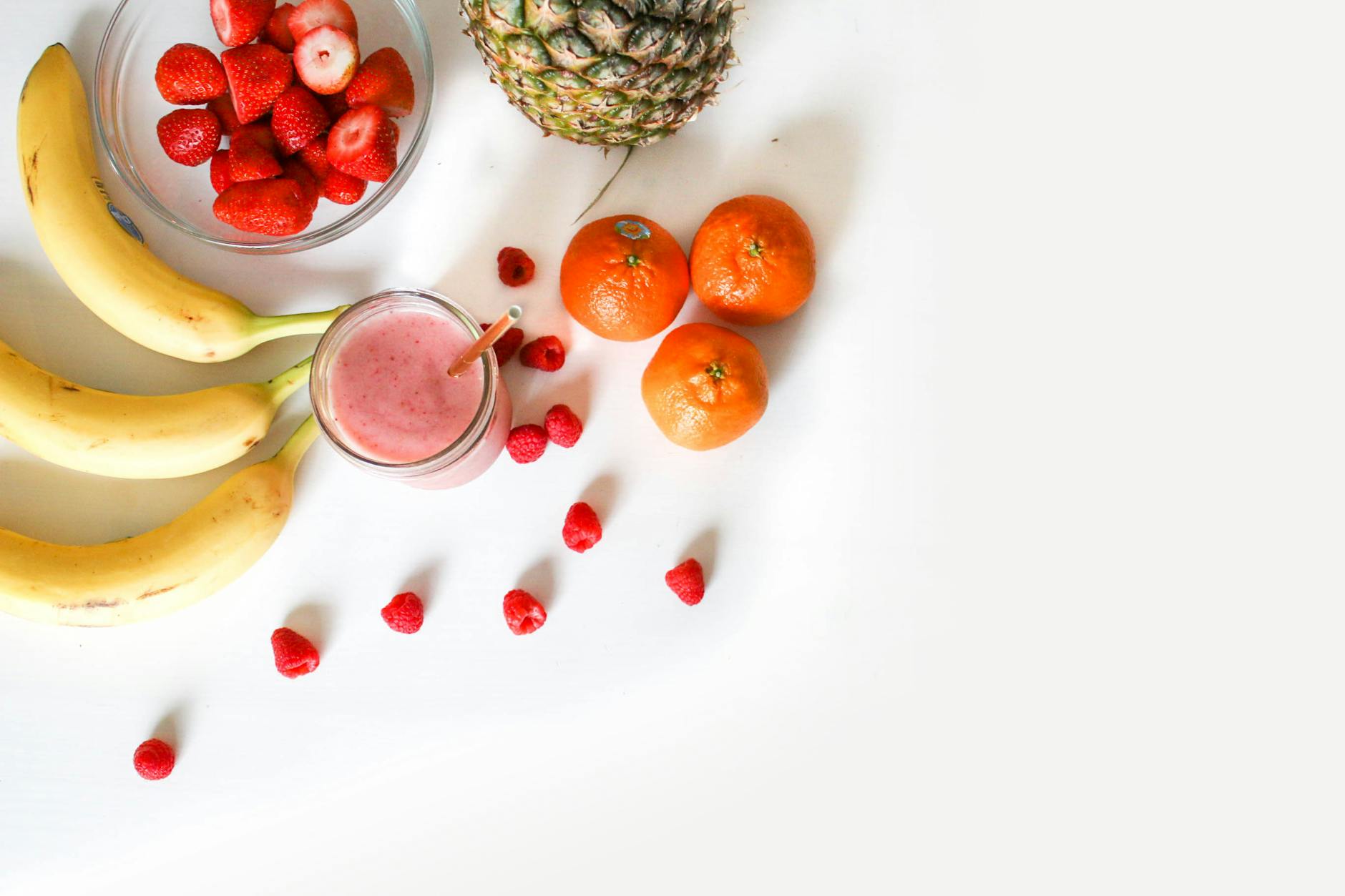 A vibrant flat lay of tropical fruits including bananas, strawberries, and a smoothie on a white background. - healthy smoothie recipe