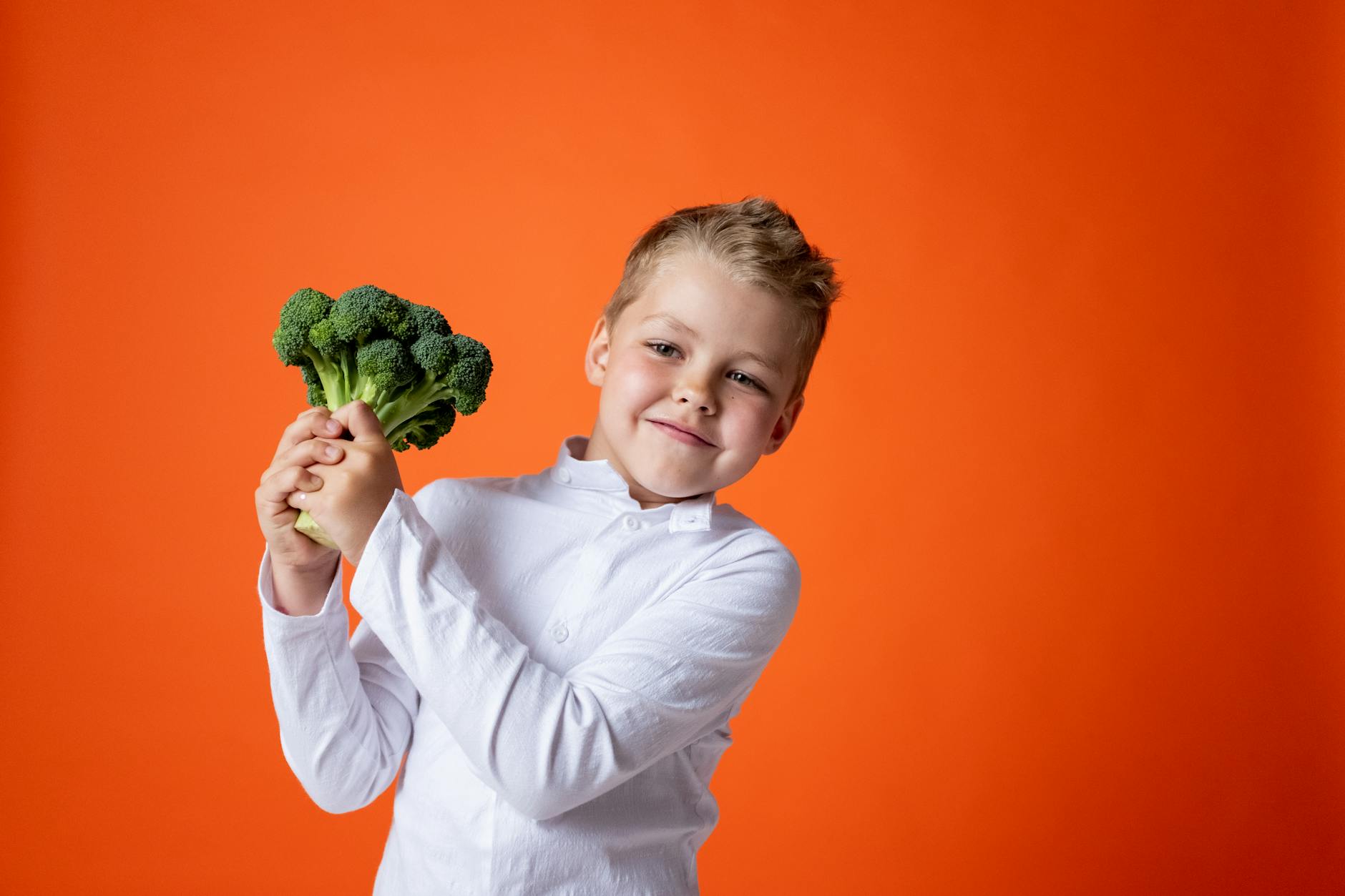 Cheerful young boy holding broccoli with a bright orange background. - healthy snacks for kids