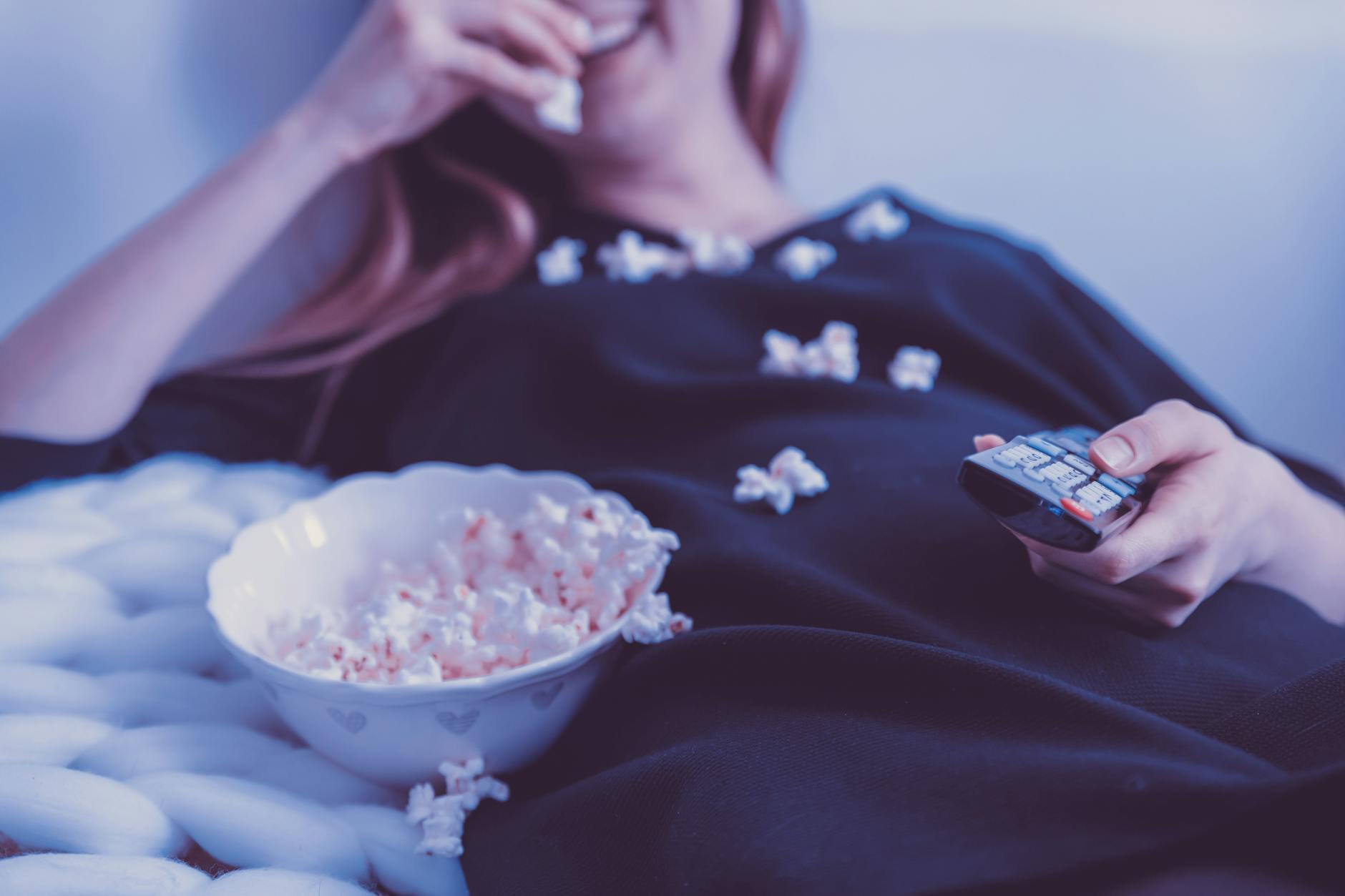 Woman relaxing on couch eating popcorn while watching TV, holding remote control, indoors. - healthy snacks near me